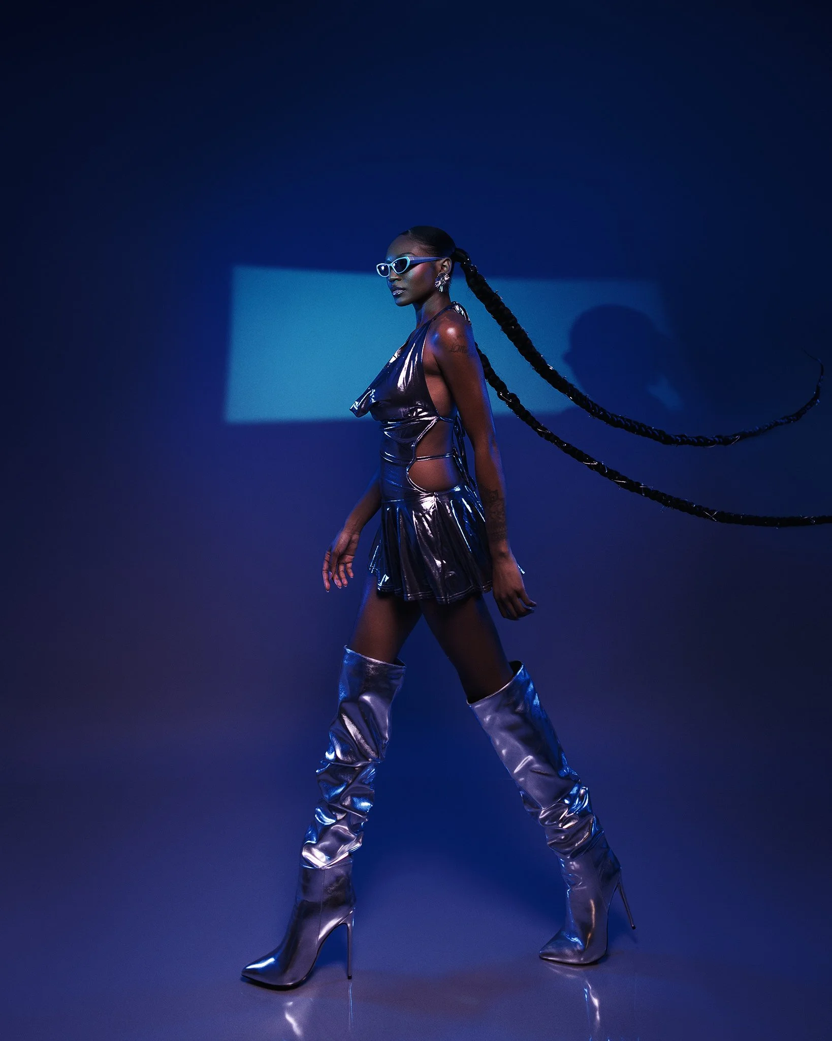 A woman with long braided hair, wearing silver high-heeled boots, sunglasses, a metallic dress with cutouts, walking against a dark blue background with a rectangular light. She has a confident stance and jewelry, and her shadow is cast on the wall b