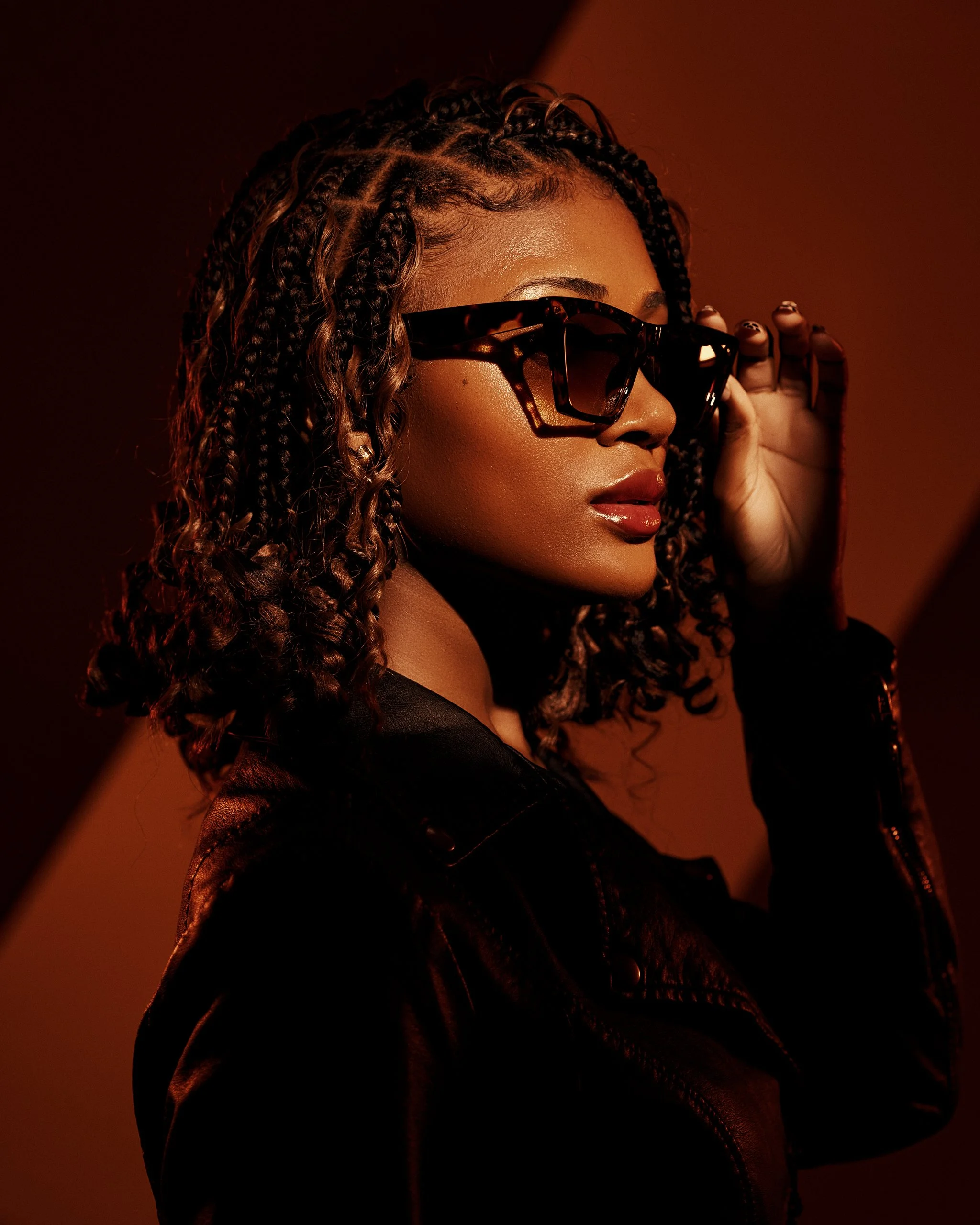 A woman with braided hair wearing dark sunglasses, bold lipstick, and a black leather jacket, posing against a warm, gradient background.