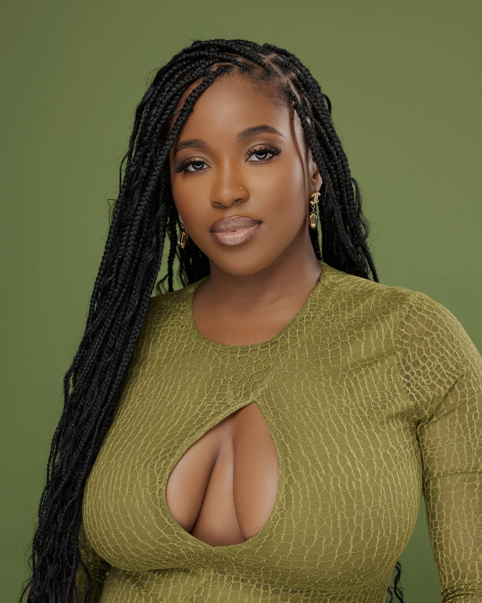 Portrait of a young woman with long braided hair, wearing a green textured top with a keyhole cutout, and gold earrings, against a green background.