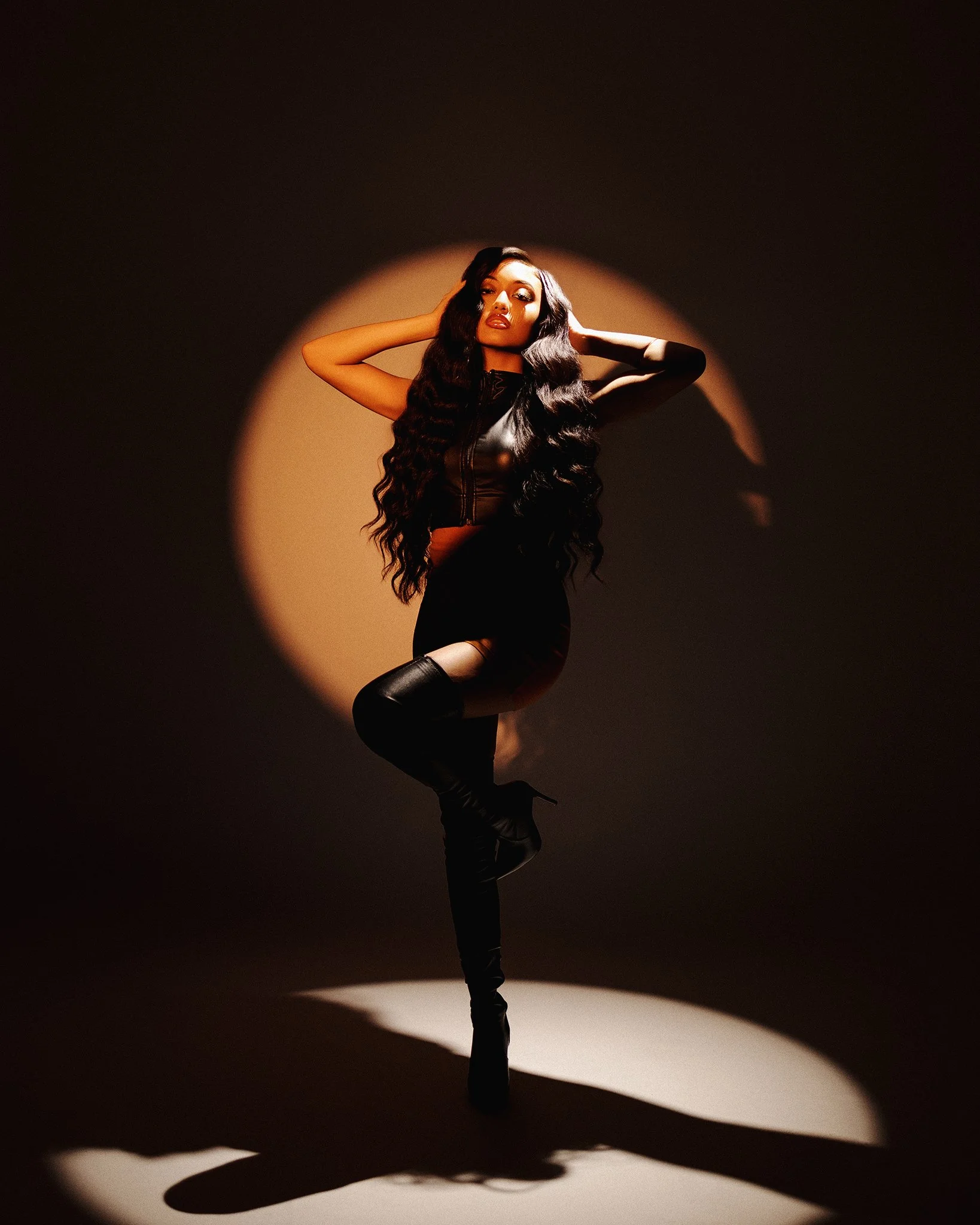 A woman with long black hair, wearing a black leather top, a short black skirt, and thigh-high black boots, striking a pose in front of a spotlight with a shadow on the wall behind her.