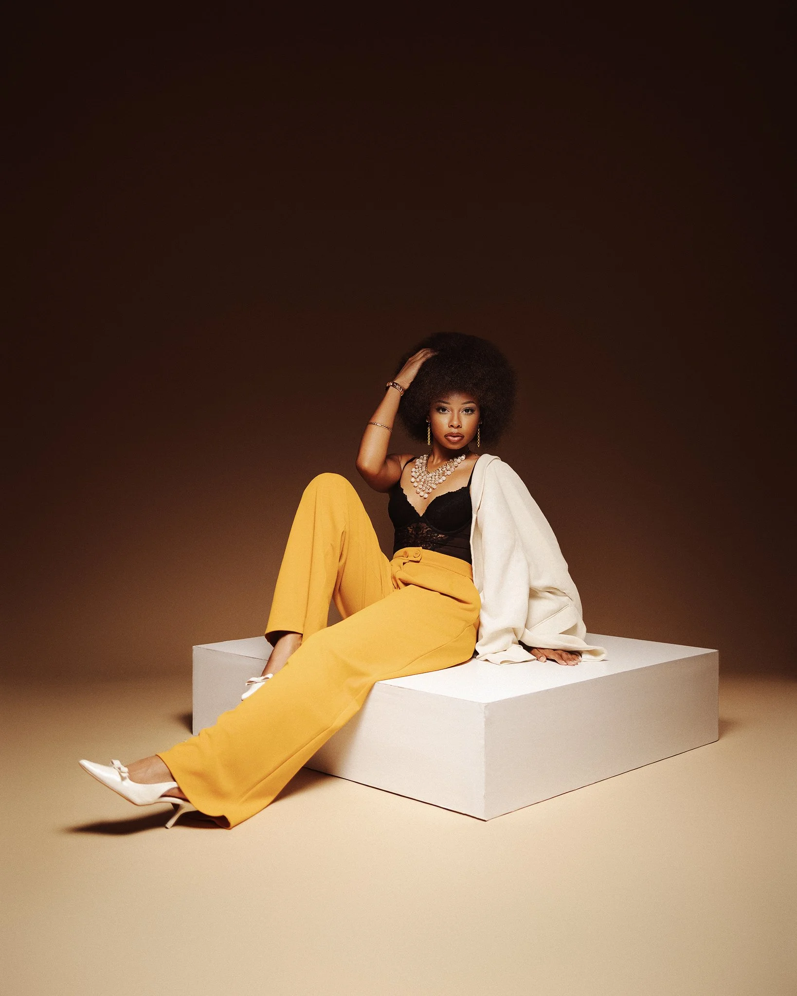 A woman with an afro hairstyle sitting on a white platform against a brown background. She is wearing yellow pants, a black top, a white jacket draped over her arm, white heels, and jewelry including a necklace and earrings.