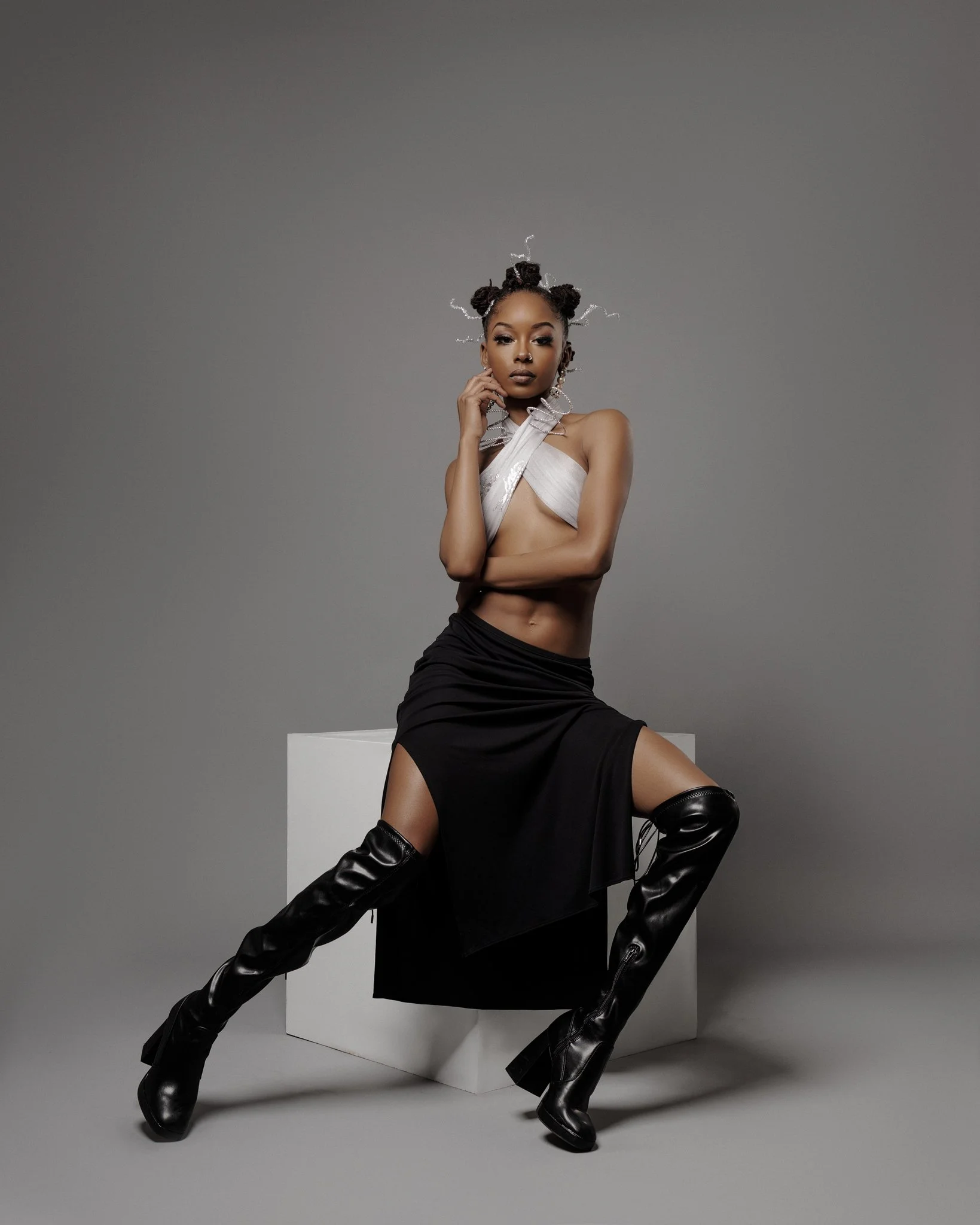 Fashion model with styled hair, wearing a silver top, black skirt with high slit, and over-the-knee black leather boots, posing with crossed legs and hand on chin against a gray background.
