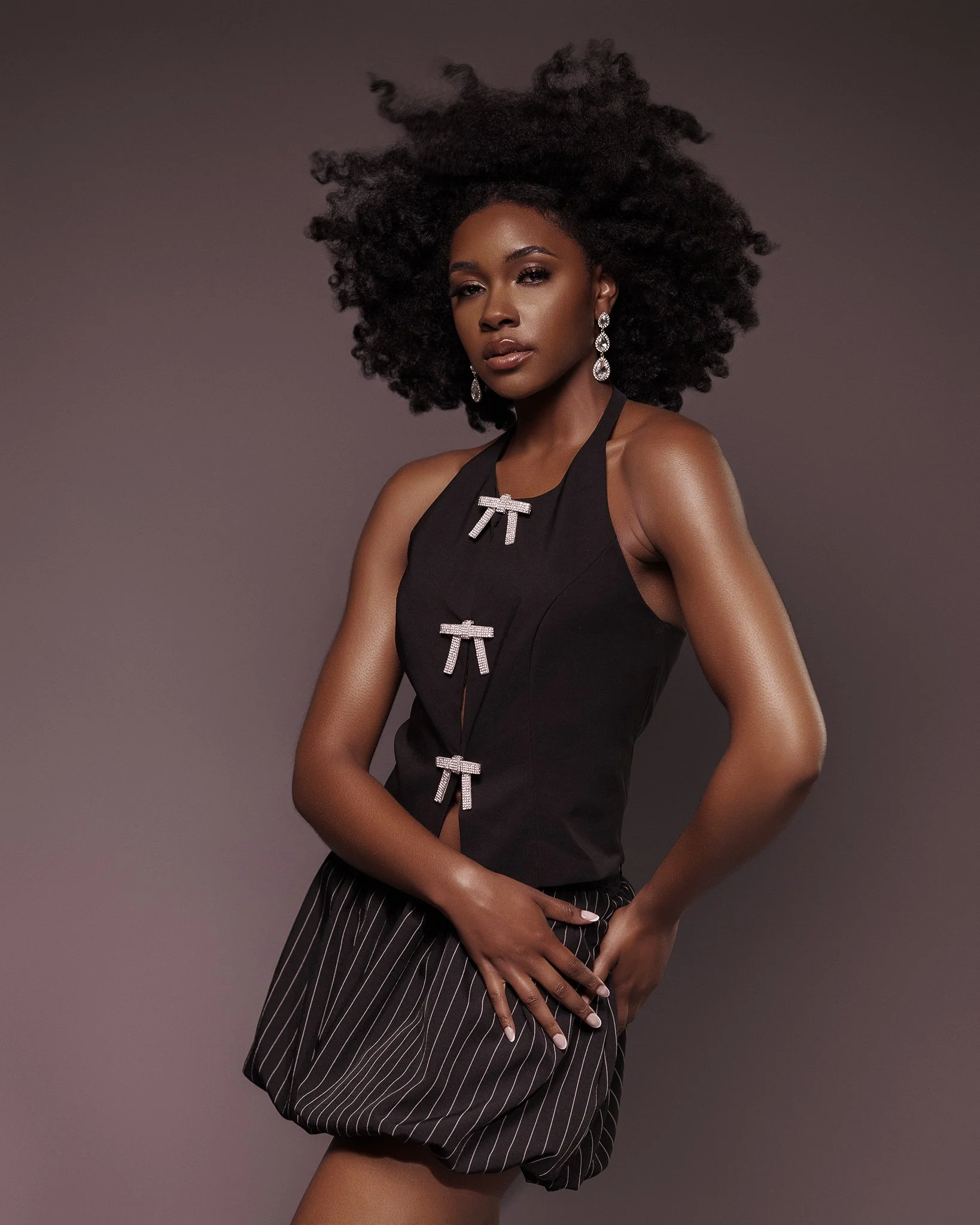 A woman with natural curly hair wearing a black sleeveless top with three rhinestone bows and striped shorts, standing against a plain background.