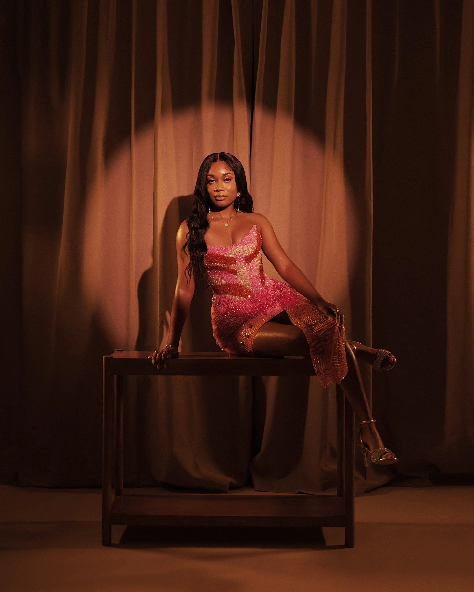 A woman in a pink and gold dress sitting on a table in front of a brown curtain
