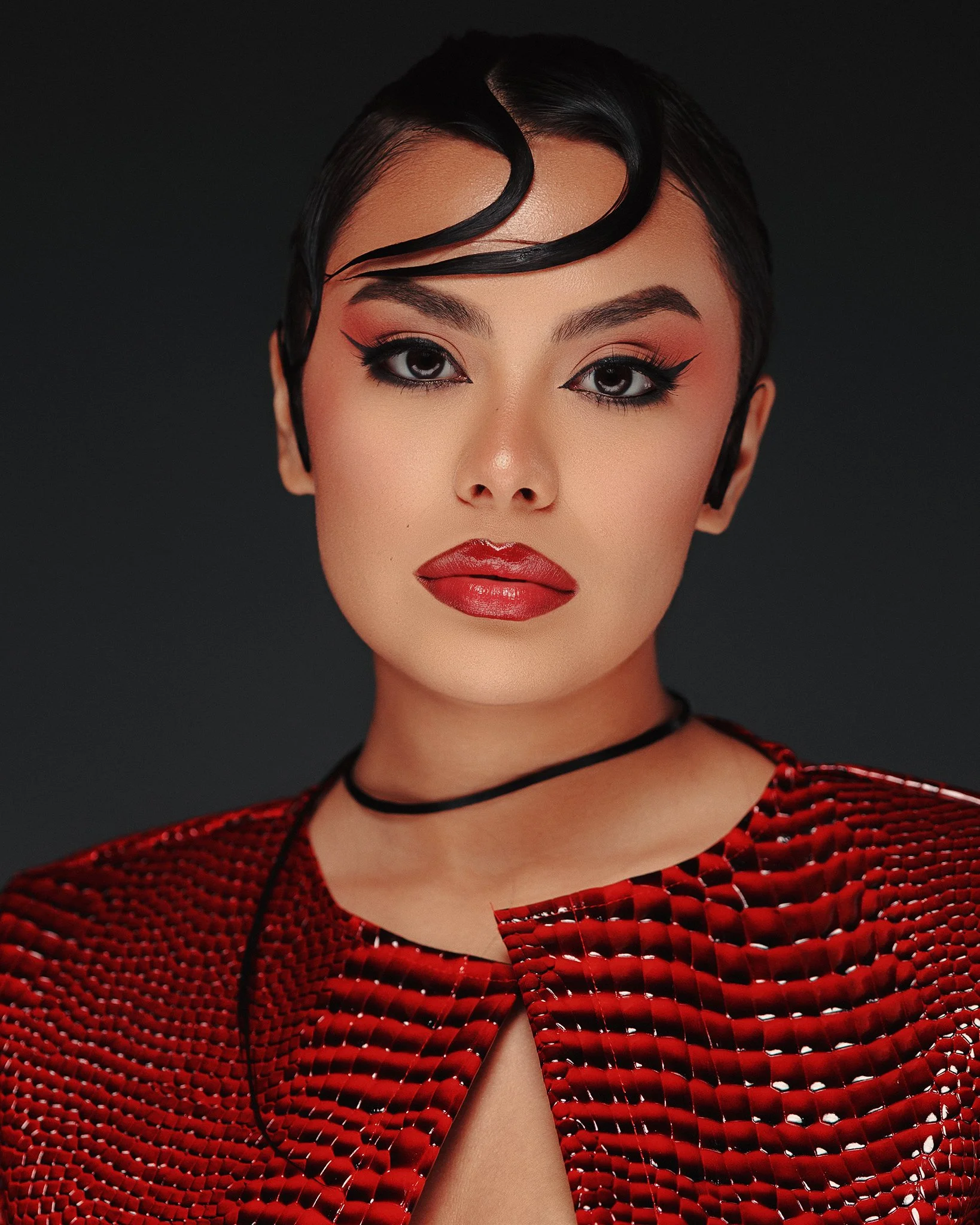 A woman with short black hair styled with a wave, bold makeup featuring winged eyeliner and red lipstick, wearing a red textured top with a keyhole neckline and a black choker.