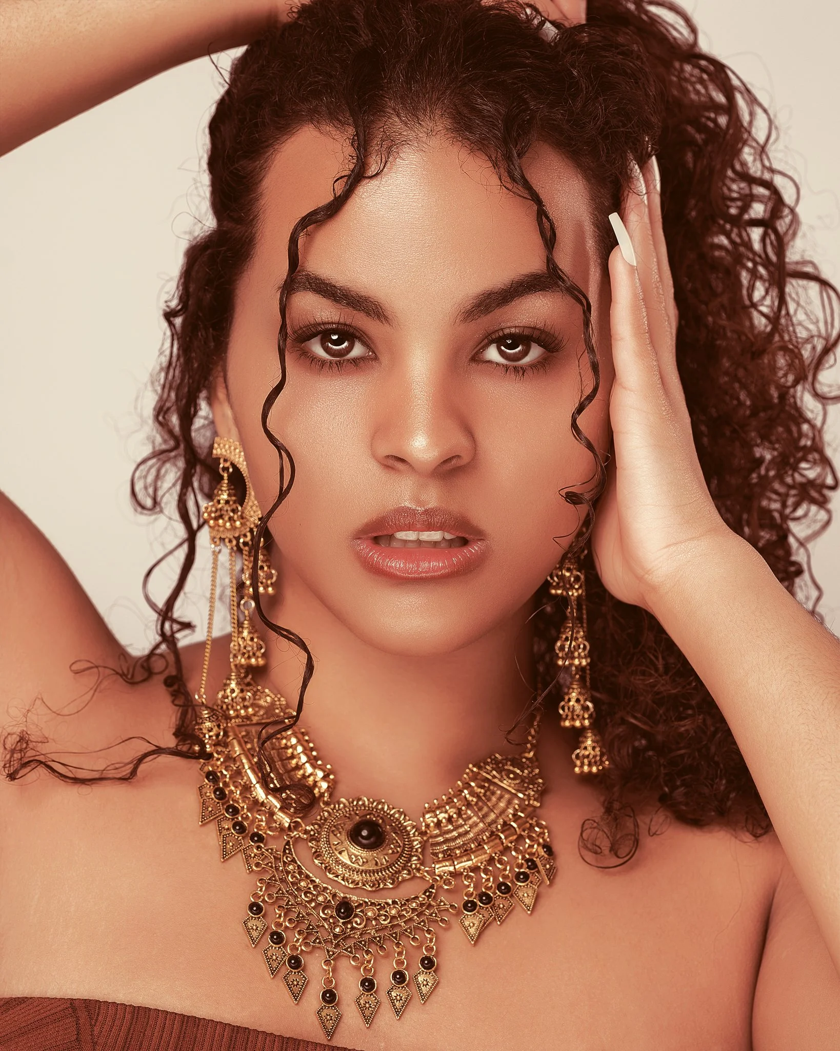 Close-up of a woman with curly hair wearing intricate gold jewelry and a strapless top, with a neutral background.