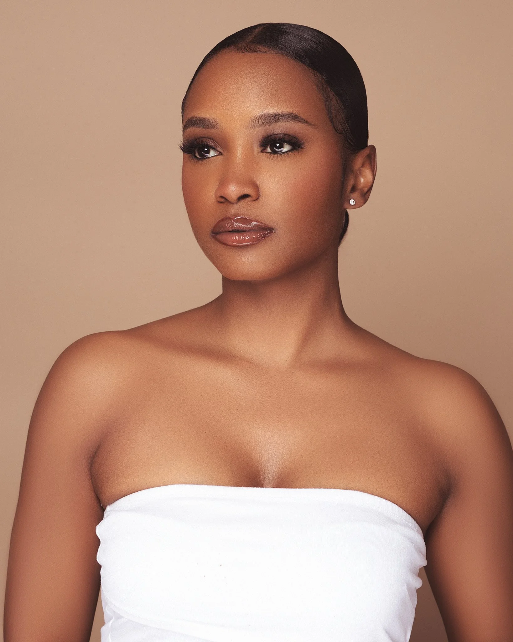 A woman with dark hair in a sleek hairstyle, makeup, and a strapless white top, looking slightly to the side against a beige background.