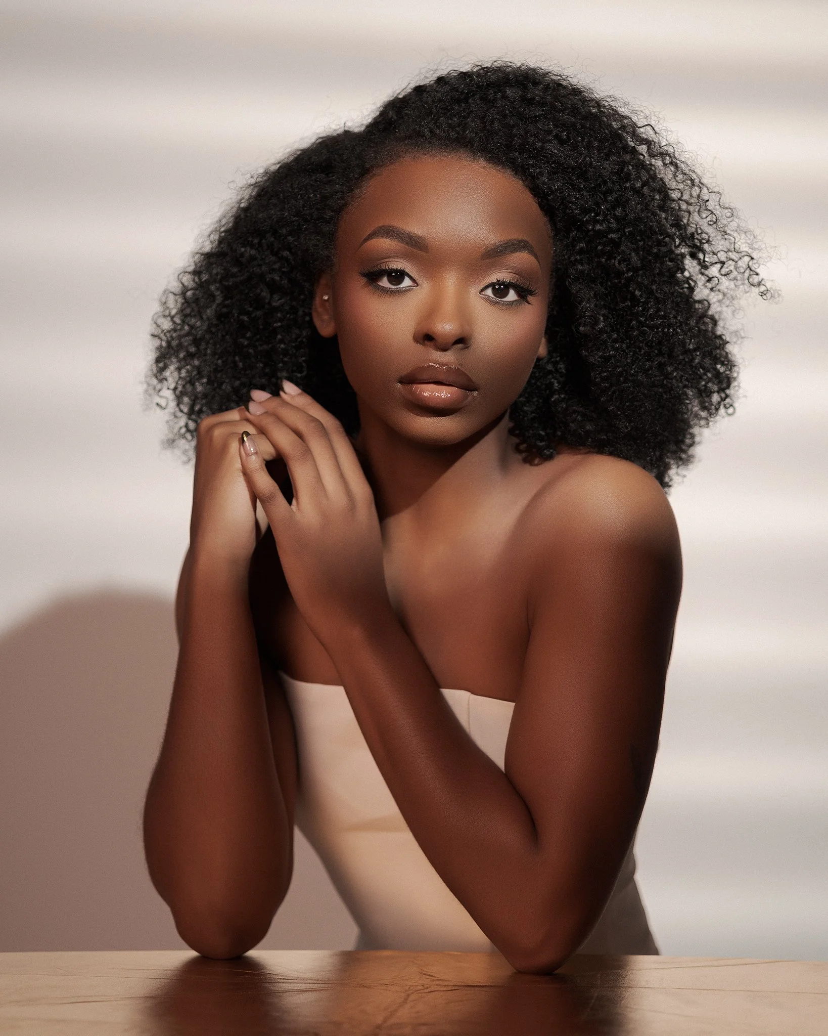 portrait of a young woman with curly black hair, wearing a strapless beige top, sitting at a wooden surface against a neutral background.