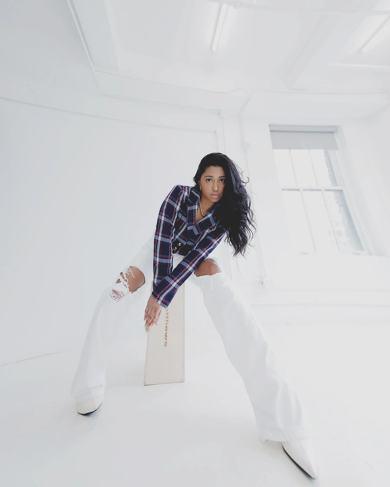 Young woman with long black hair wearing ripped white jeans, a plaid shirt, and white shoes, standing in a bright white room with large windows.