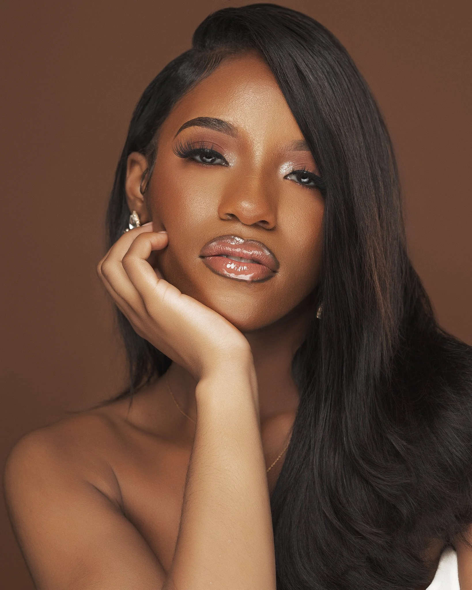 Close-up portrait of a woman with long black hair, wearing makeup with shimmery eyeshadow, glossy lips, and earrings, resting her face on her hand, against a brown background.