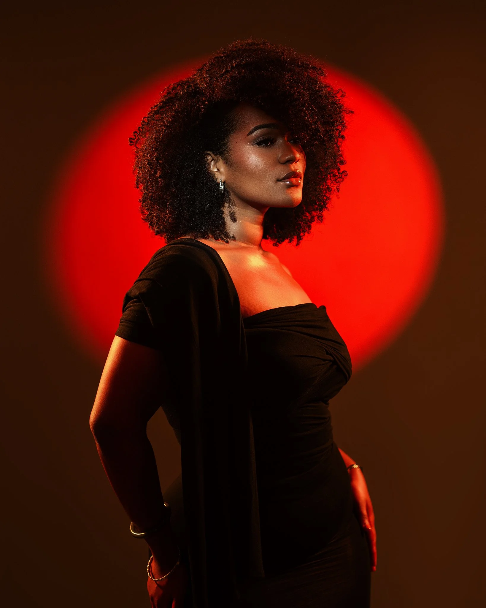 A woman with curly black hair and a black off-shoulder dress poses against a dark background with a red circular glow behind her.