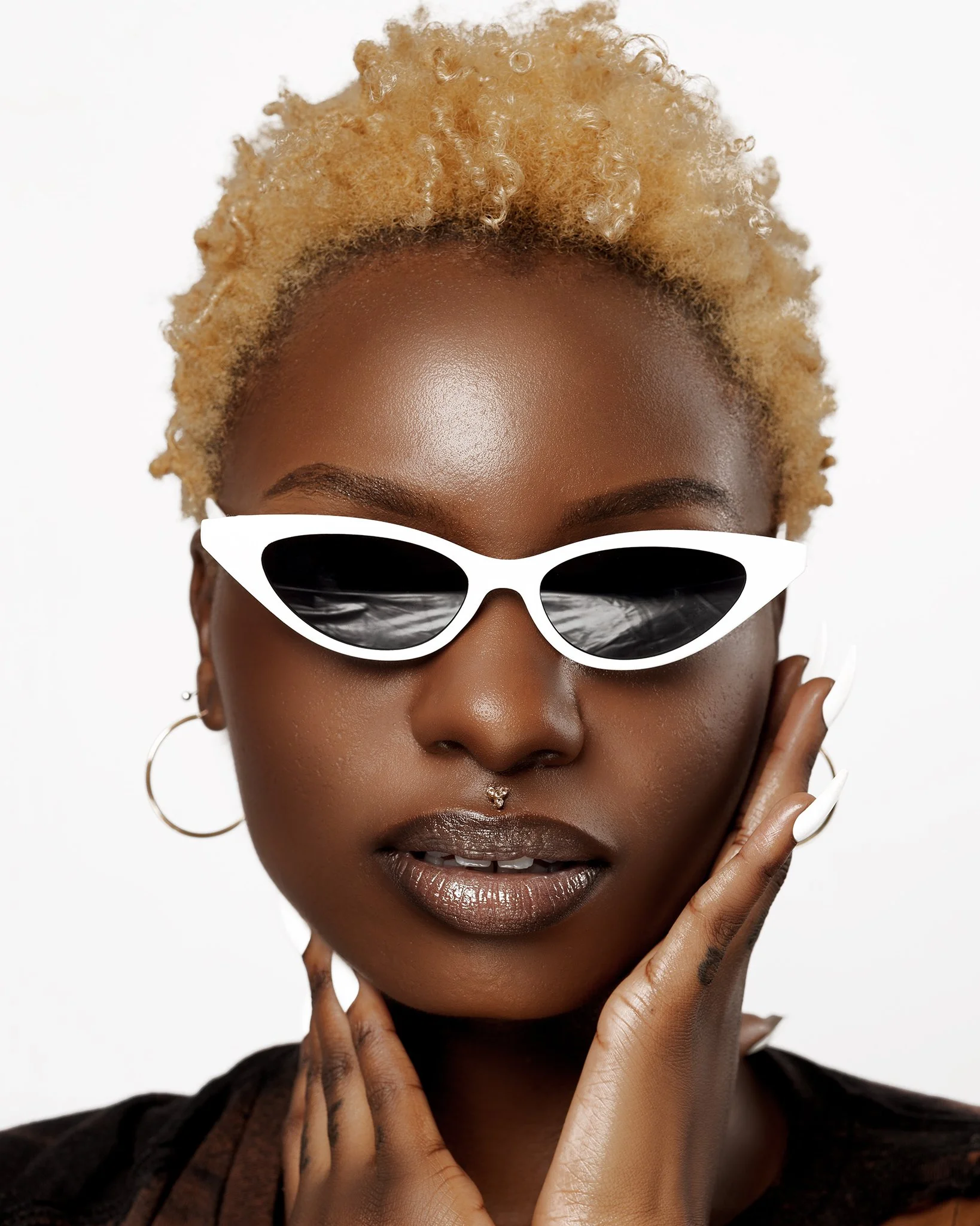 Close-up portrait of a woman with dark skin and blonde curly hair, wearing white, cat-eye sunglasses, hoop earrings, and a septum piercing, touching her face gently.