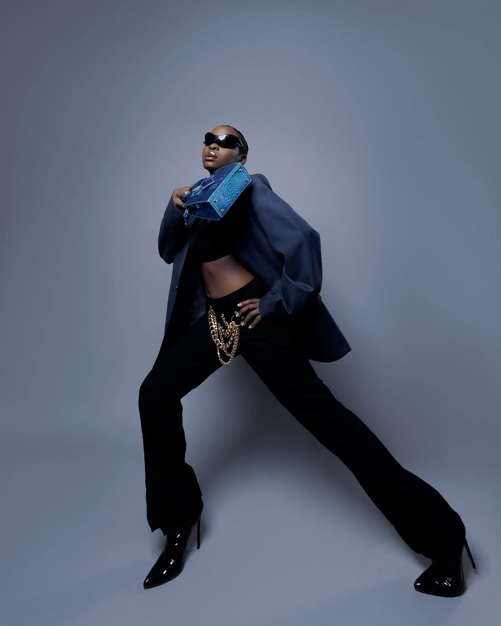 A woman striking a pose against a plain background, wearing sunglasses, a dark oversized blazer draped over her shoulders, black high-heeled boots, and accessorized with multiple gold chains and a small blue purse.