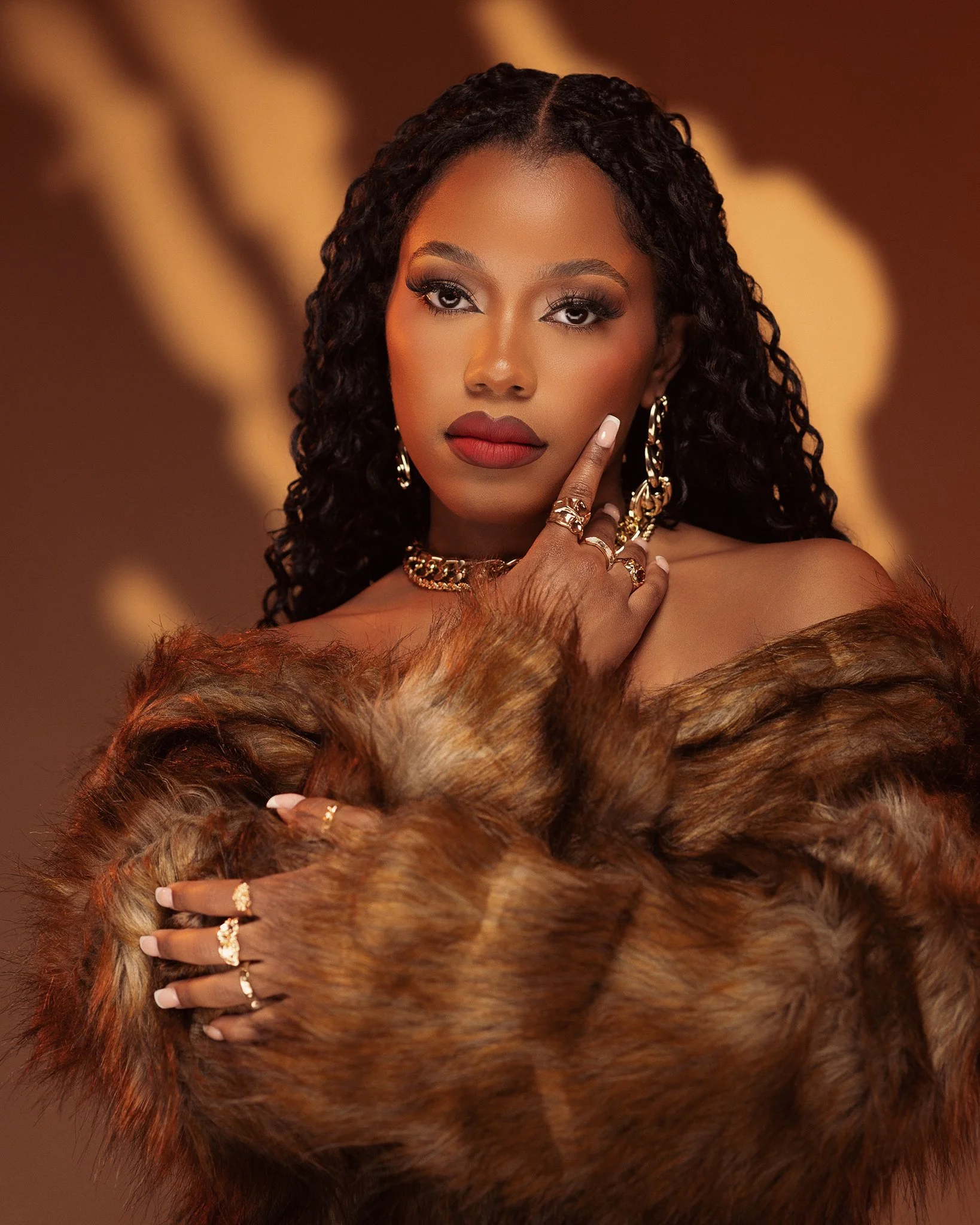 A woman with dark curly hair, wearing red lipstick and gold jewelry, poses with her hand near her face, draped in a fur coat.