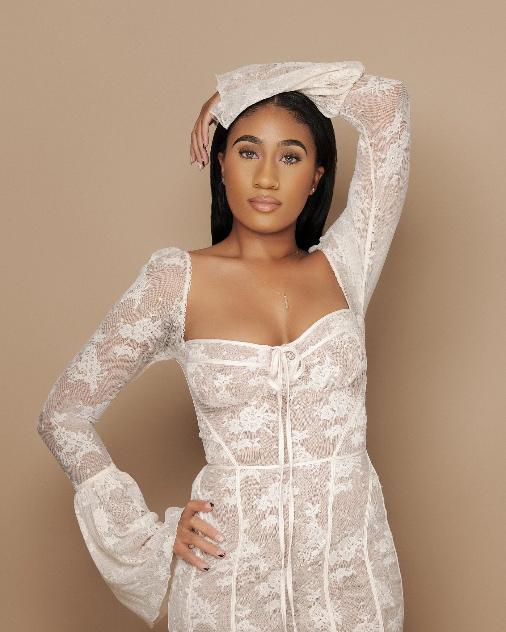 A woman with dark hair and light makeup wearing a white lace dress with long sleeves, standing against a neutral background.
