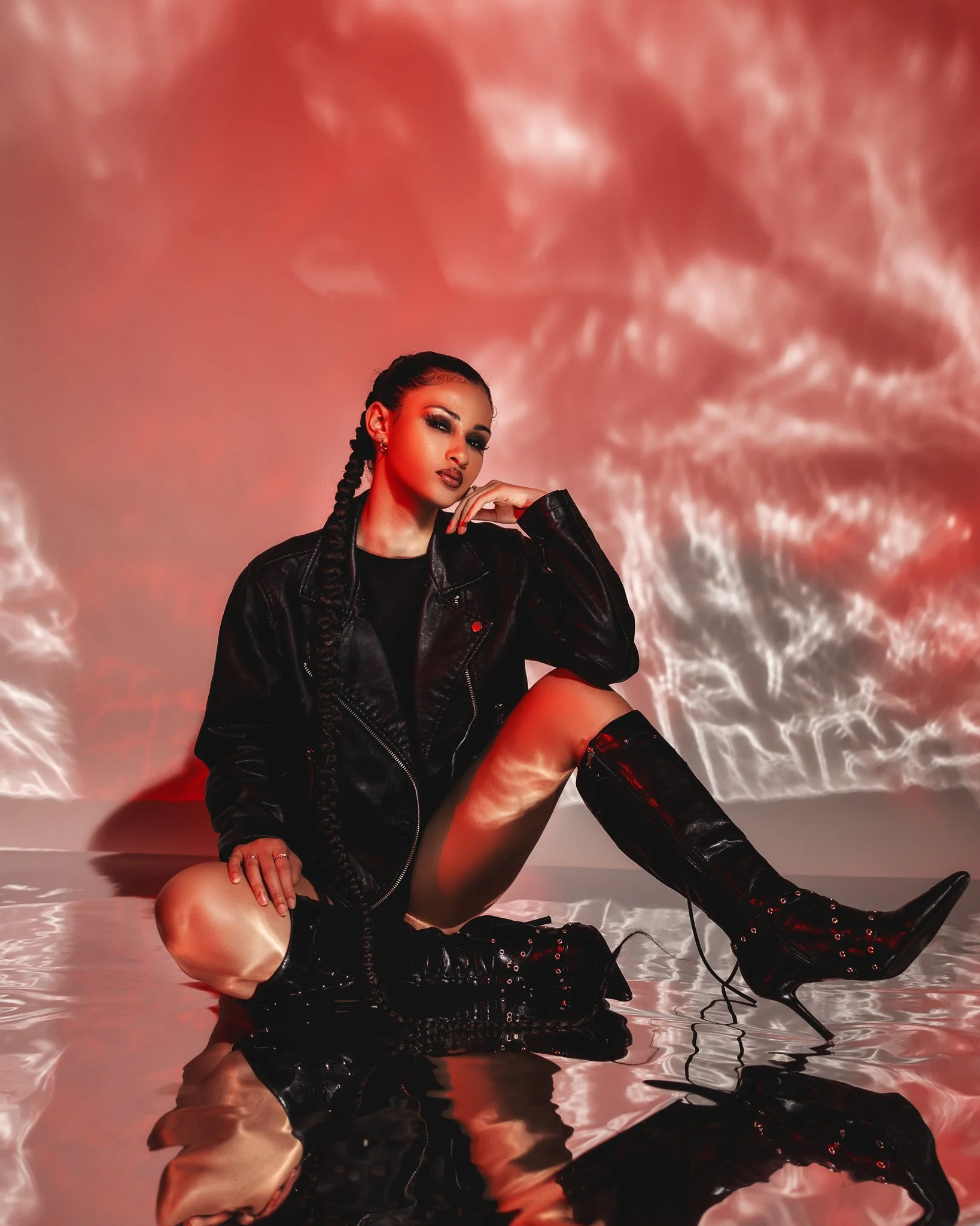A woman with braided hair, dark makeup, and black clothing sits on a reflective surface in front of a red textured background, with her foot on the ground and her hand resting near her face.