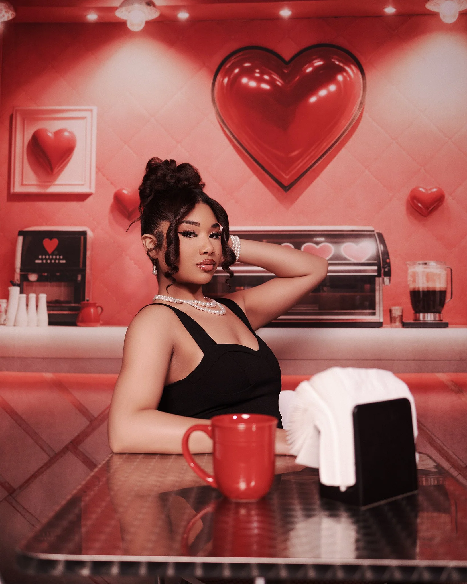A woman in a black dress sitting at a table in a diner with a pink and red heart-themed decor background.