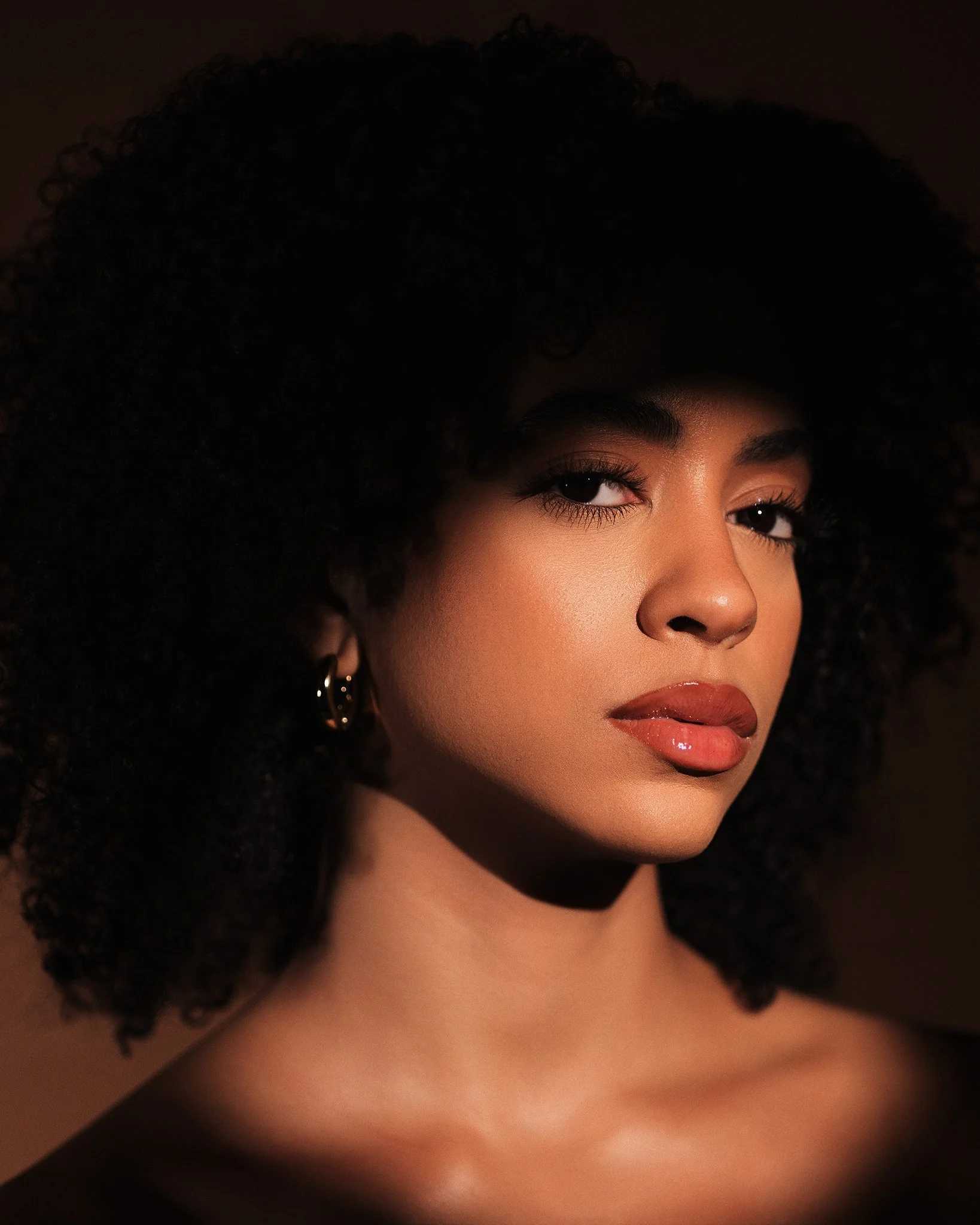 Close-up portrait of a woman with dark curly hair, wearing hoop earrings, with a confident expression, slightly tilted head, warm skin tone, and makeup including glossy lips and subtle eye makeup.