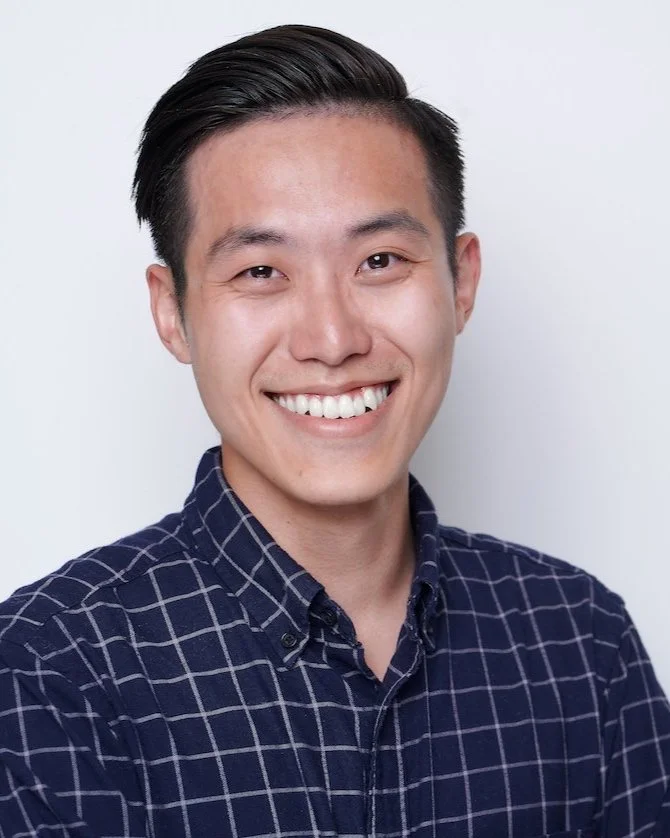 A smiling Asian man with dark hair styled to the side, wearing a navy blue checkered shirt, against a plain white background.