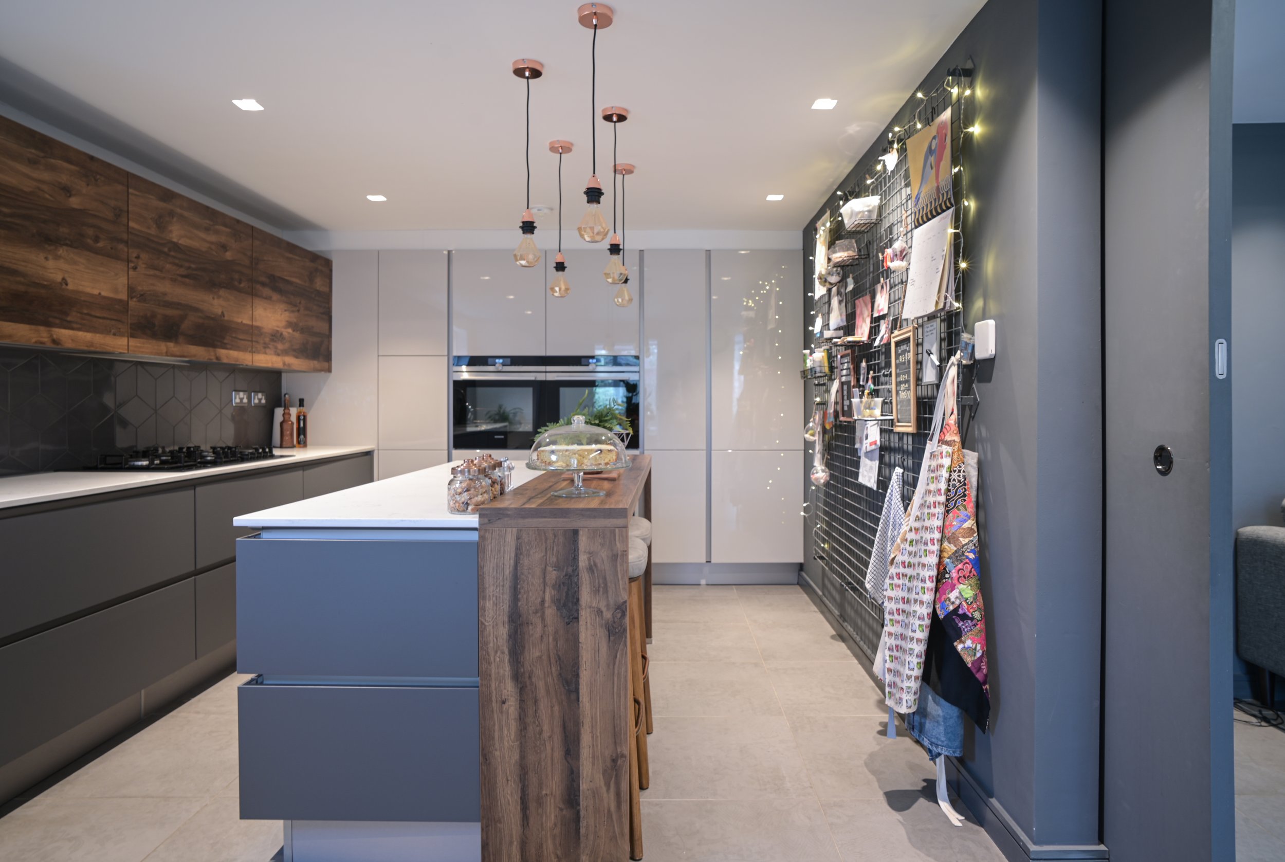 Modern kitchen with gray cabinets, a white countertop island, and wooden accents. Features pendant lighting, wall-mounted storage grid with kitchen items, and a small table with chairs.