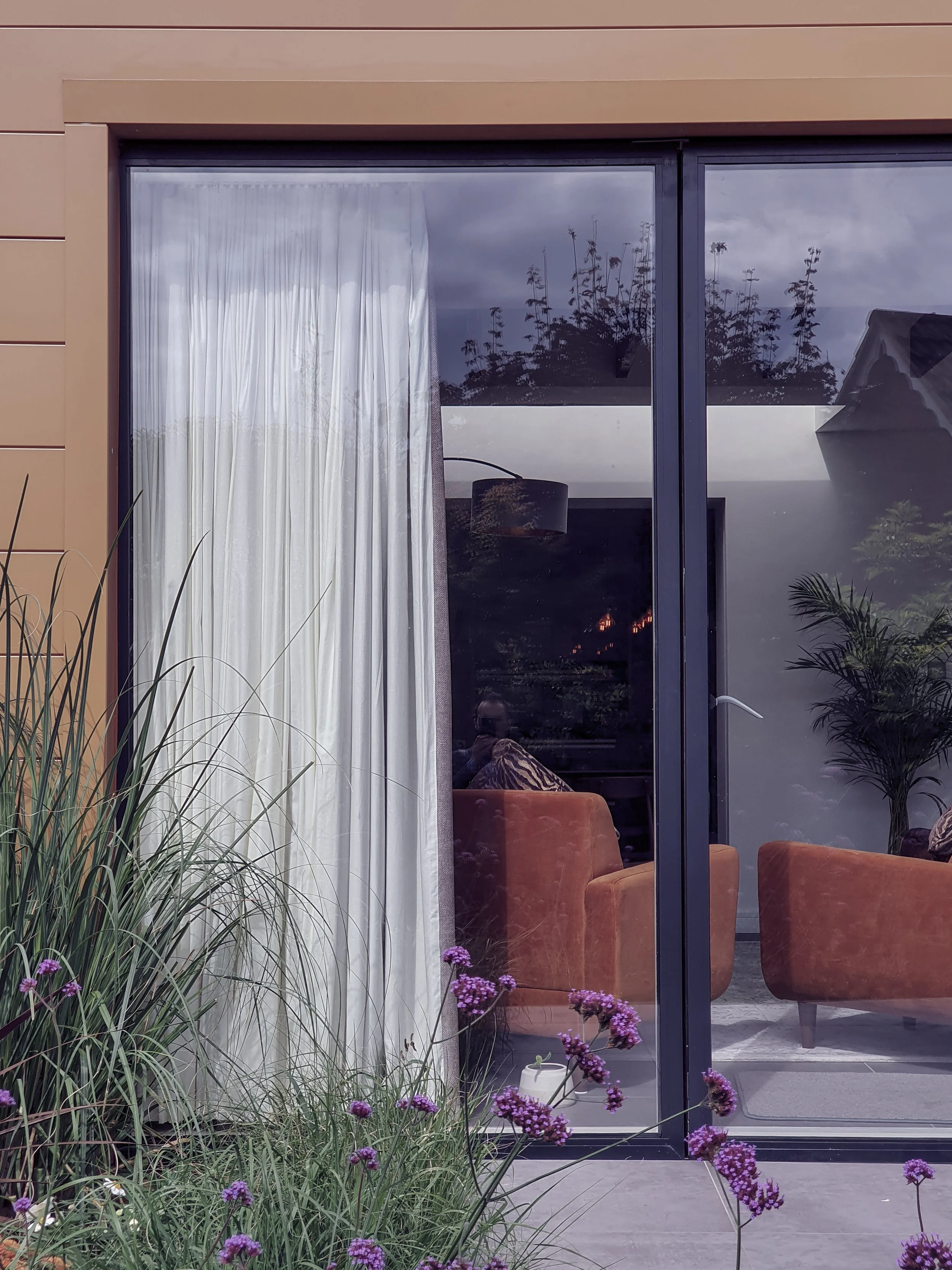 View of a living room seen through a large glass sliding door with a beige exterior wall. Inside, there are orange velvet armchairs, a black ceiling lamp, and some plants. Outside, there are purple flowers, tall grass, and potted plants.