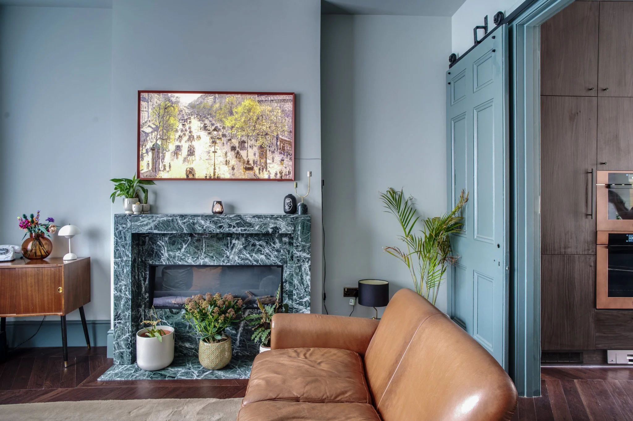 Living room with a fireplace with a marble mantel, plants, artwork, and furniture including a leather sofa.