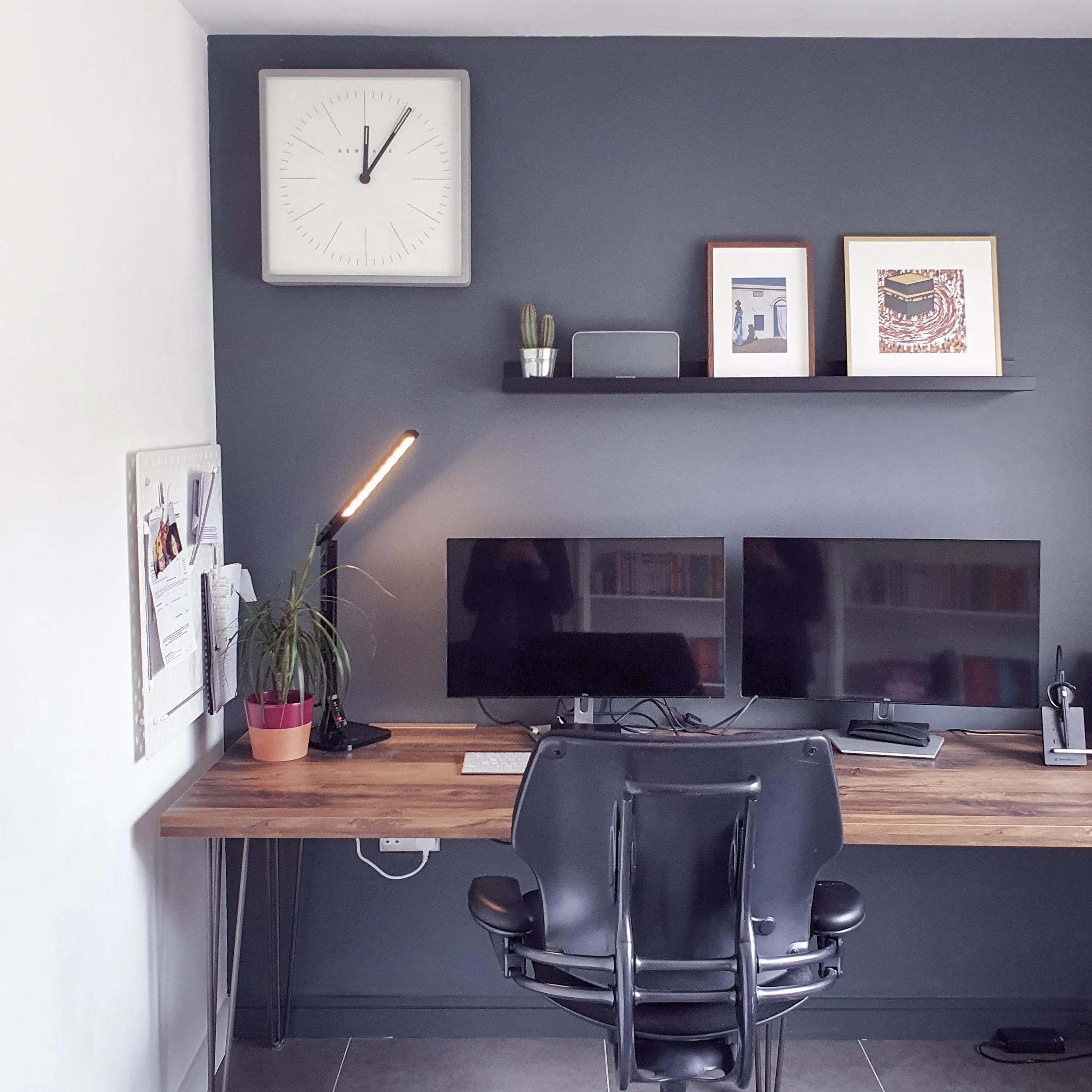 Office workspace with a dark blue accent wall, a wall clock showing 12:10, framed artwork, a potted cactus, a monitor, a keyboard, a computer mouse, and a black chair.