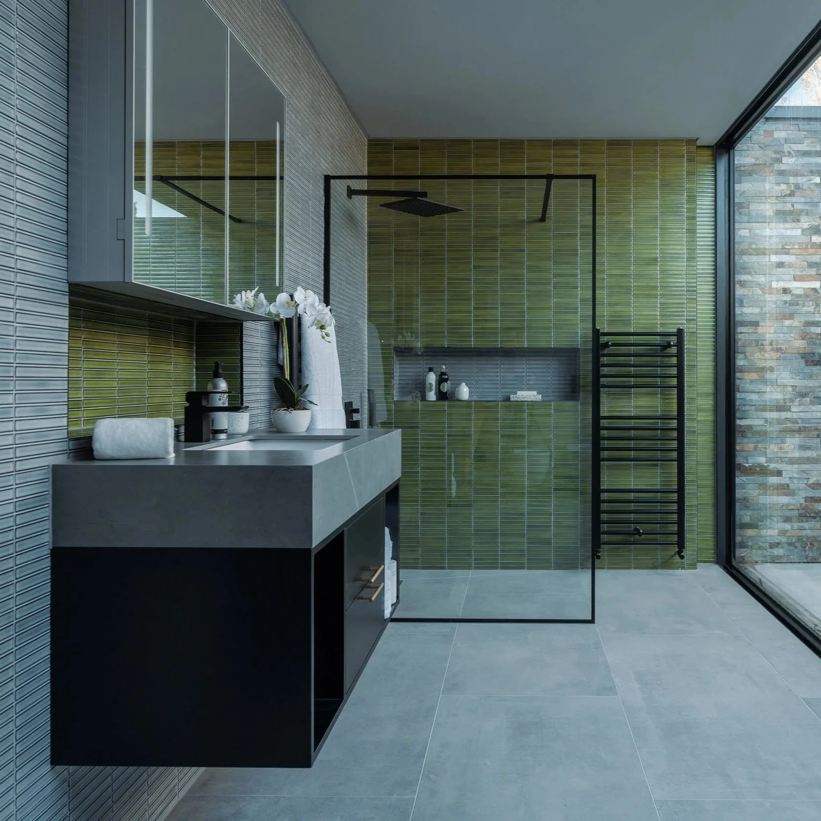 Modern bathroom with gray tiled floor, green tiled shower area, black framed glass shower door, black vanity with sink, and large window with rain view.