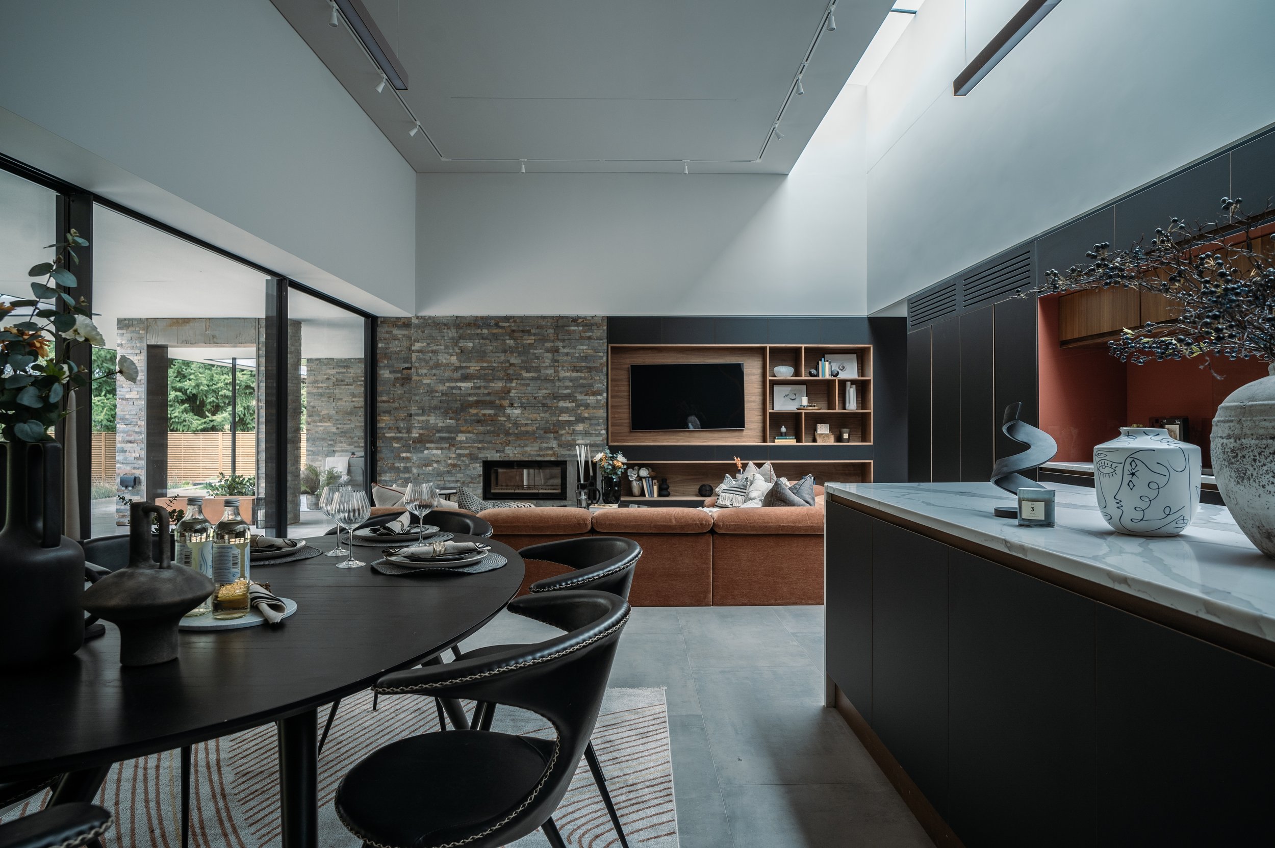 Modern open-concept living and dining area with large windows, a stone accent wall, built-in shelves with decorative items, a curved sofa, a dining table set with wine glasses and plates, and a marble kitchen countertop with decorative vases.