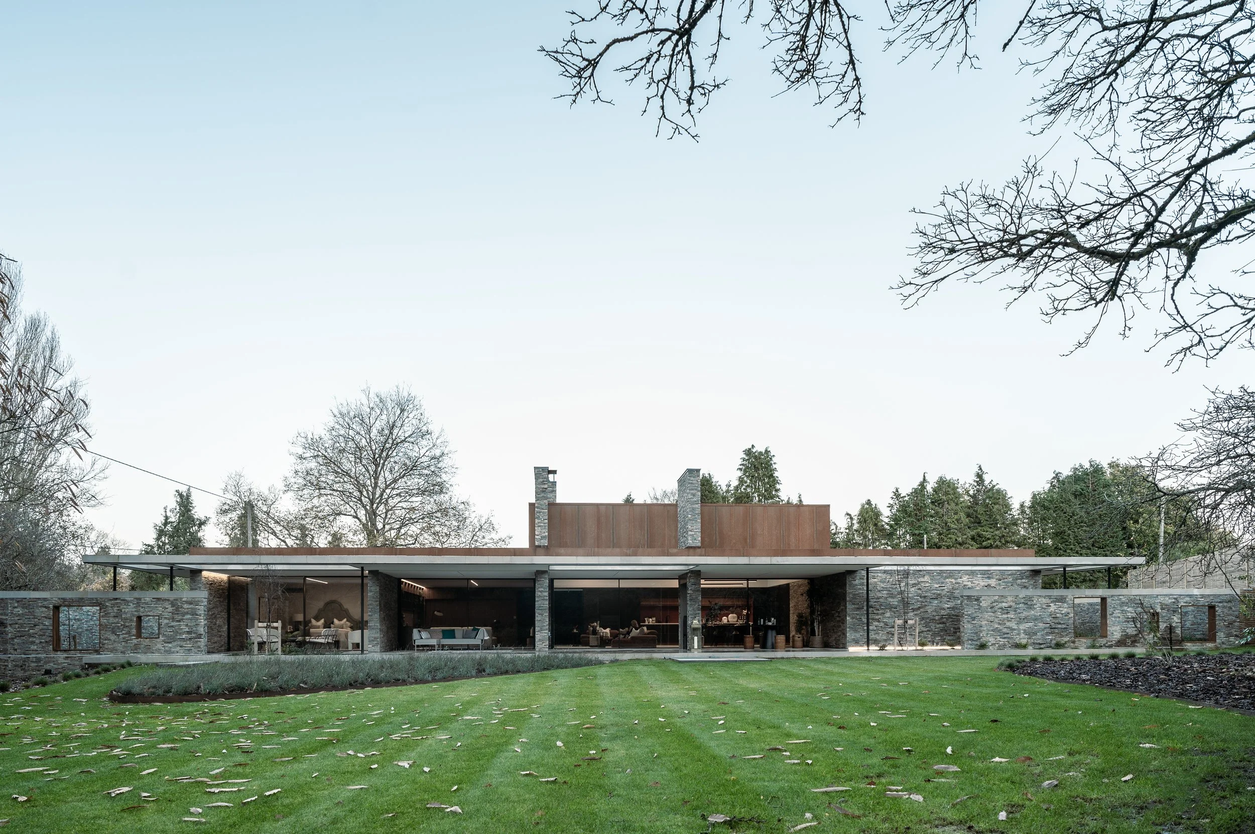 Modern house with large windows, stone walls, and a flat roof, surrounded by a green lawn with scattered fallen leaves, and tall, leafless trees against a clear sky.