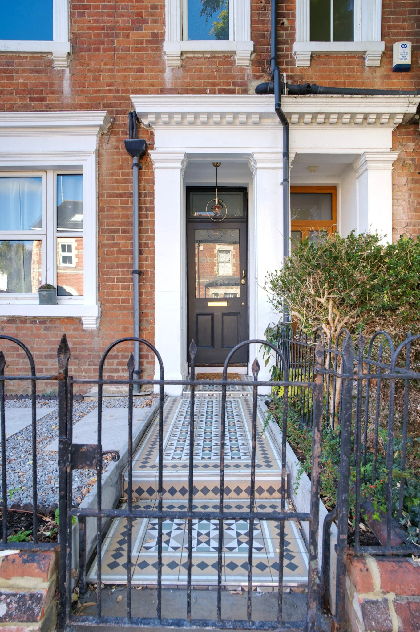 Front porch of a brick house with a decorative tiled walkway, iron gate, white door frame, and windows with white trim. Bushes and plants are on the sides.
