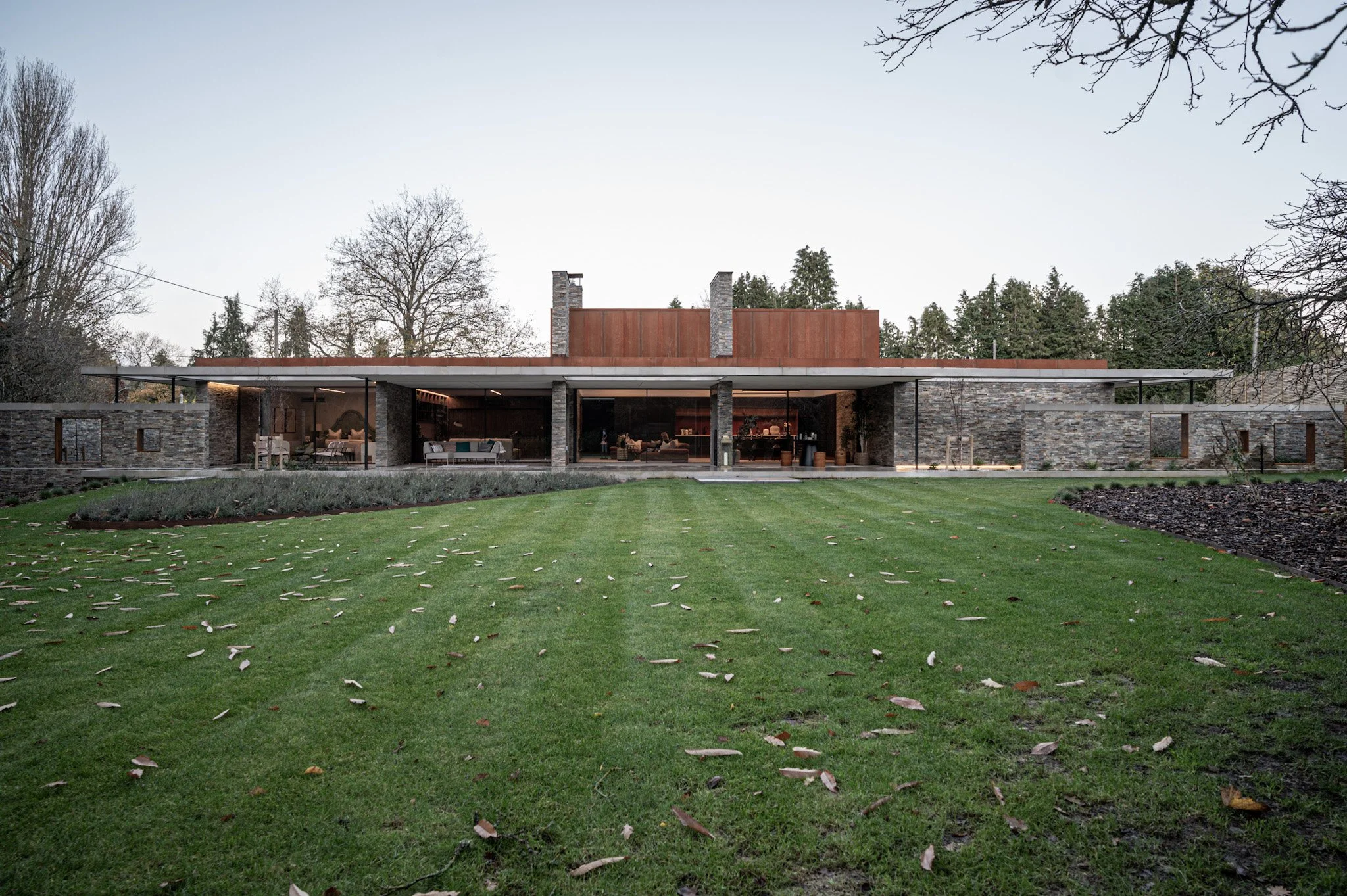 Modern house with large glass windows, stone walls, and a spacious lawn with fallen leaves, set against a backdrop of trees and a clear sky.