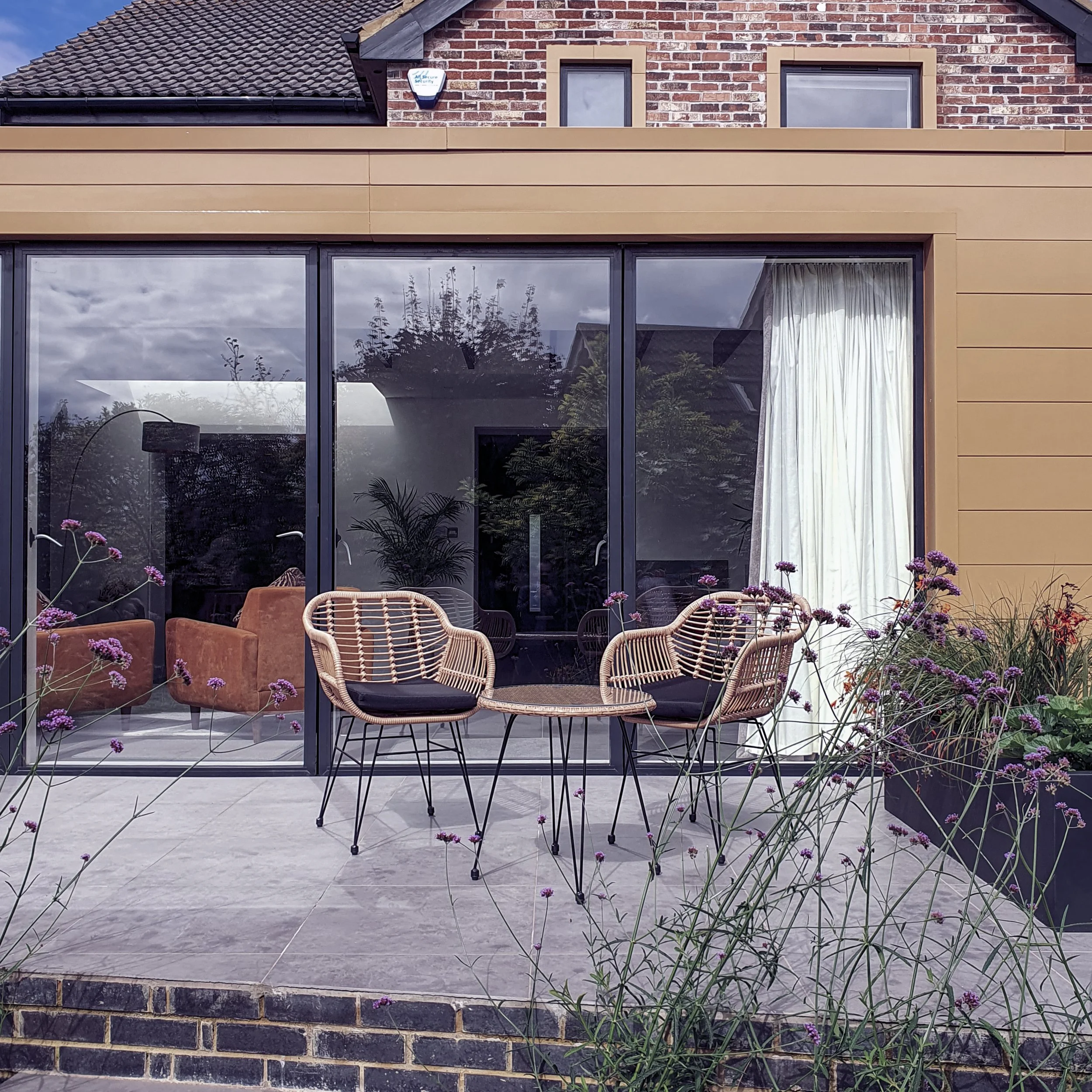 Exterior view of a modern house with a brick wall, large glass sliding doors, a tan siding, and potted plants in the garden with purple flowers.