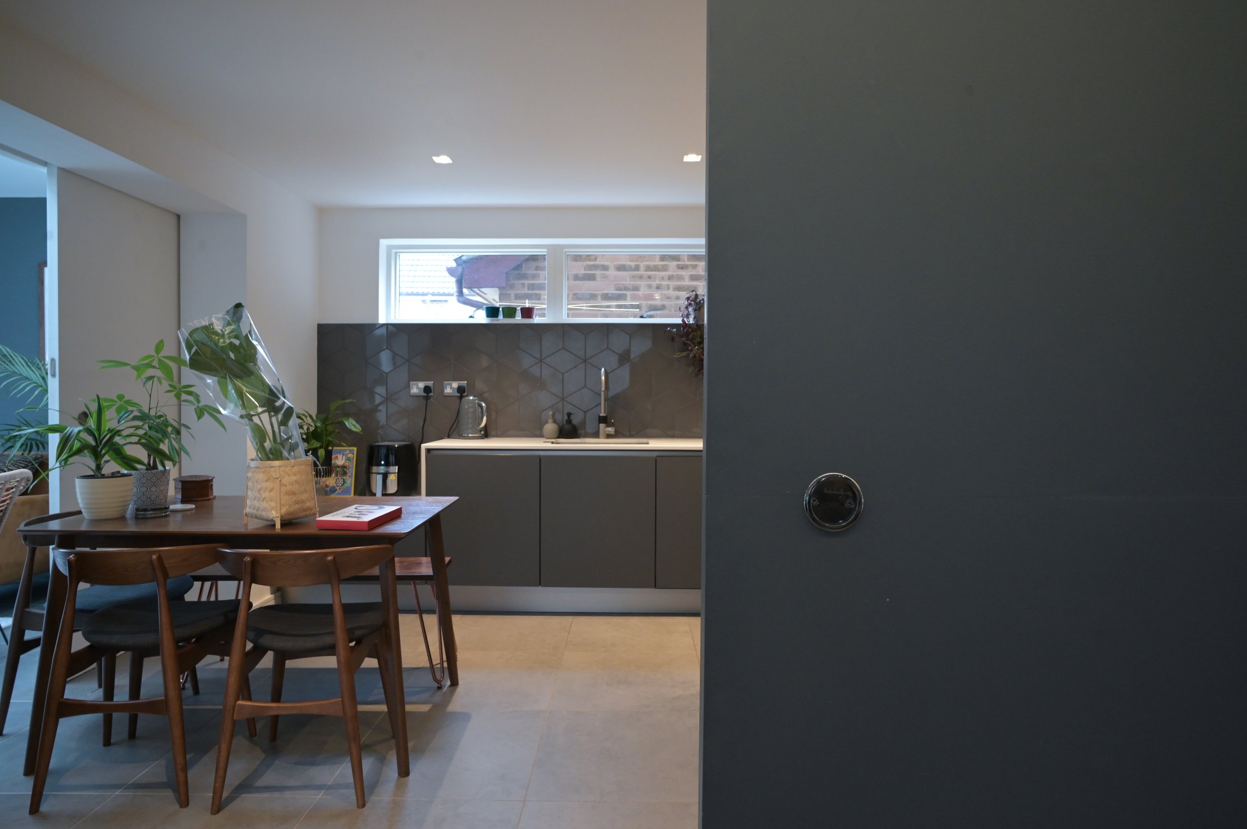 A dining area with a wooden table and chairs, green plants on the table, and a kitchen with a window, gray cabinets, and a black backsplash in the background. Part of a dark wall with a round black object is visible on the right.