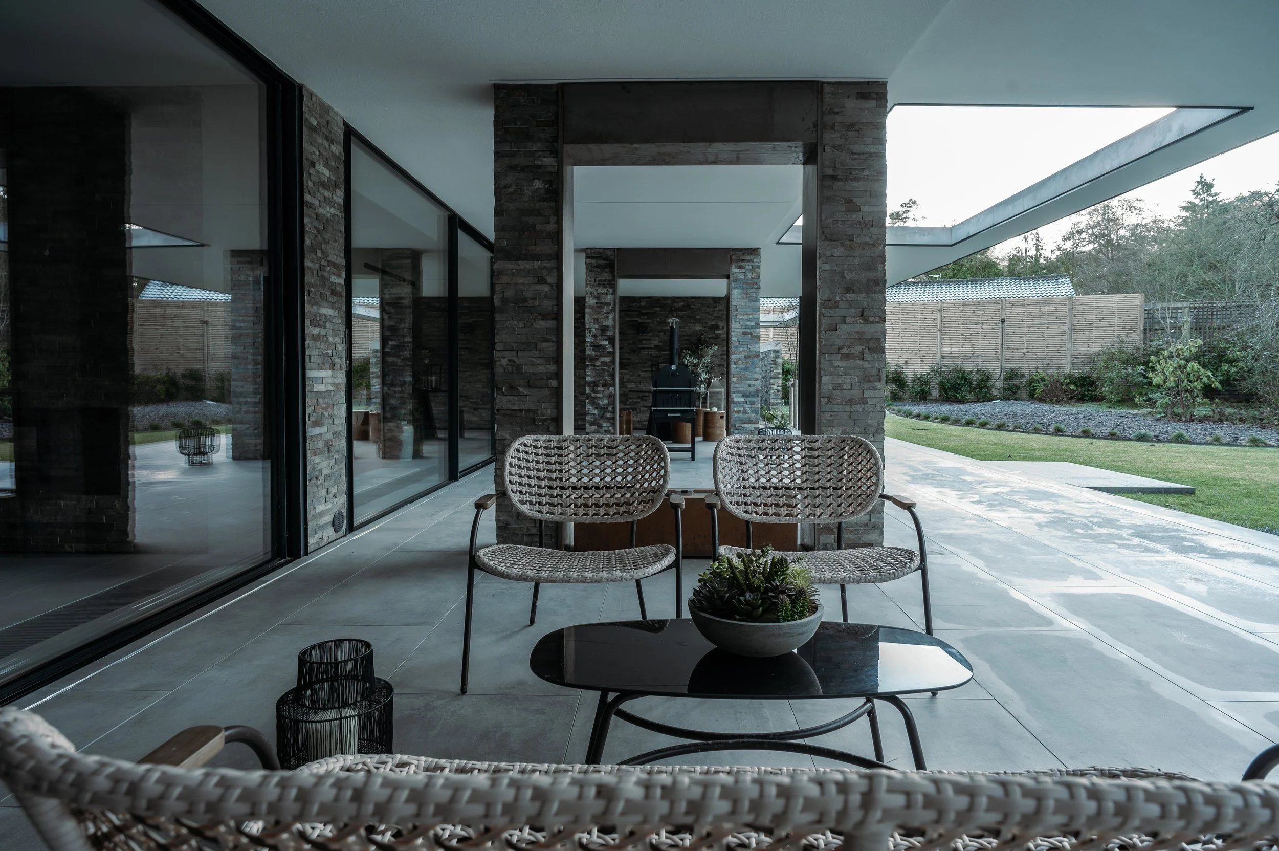 Modern house patio with outdoor seating, large sliding glass doors, and a view of a garden with trees and a fence.