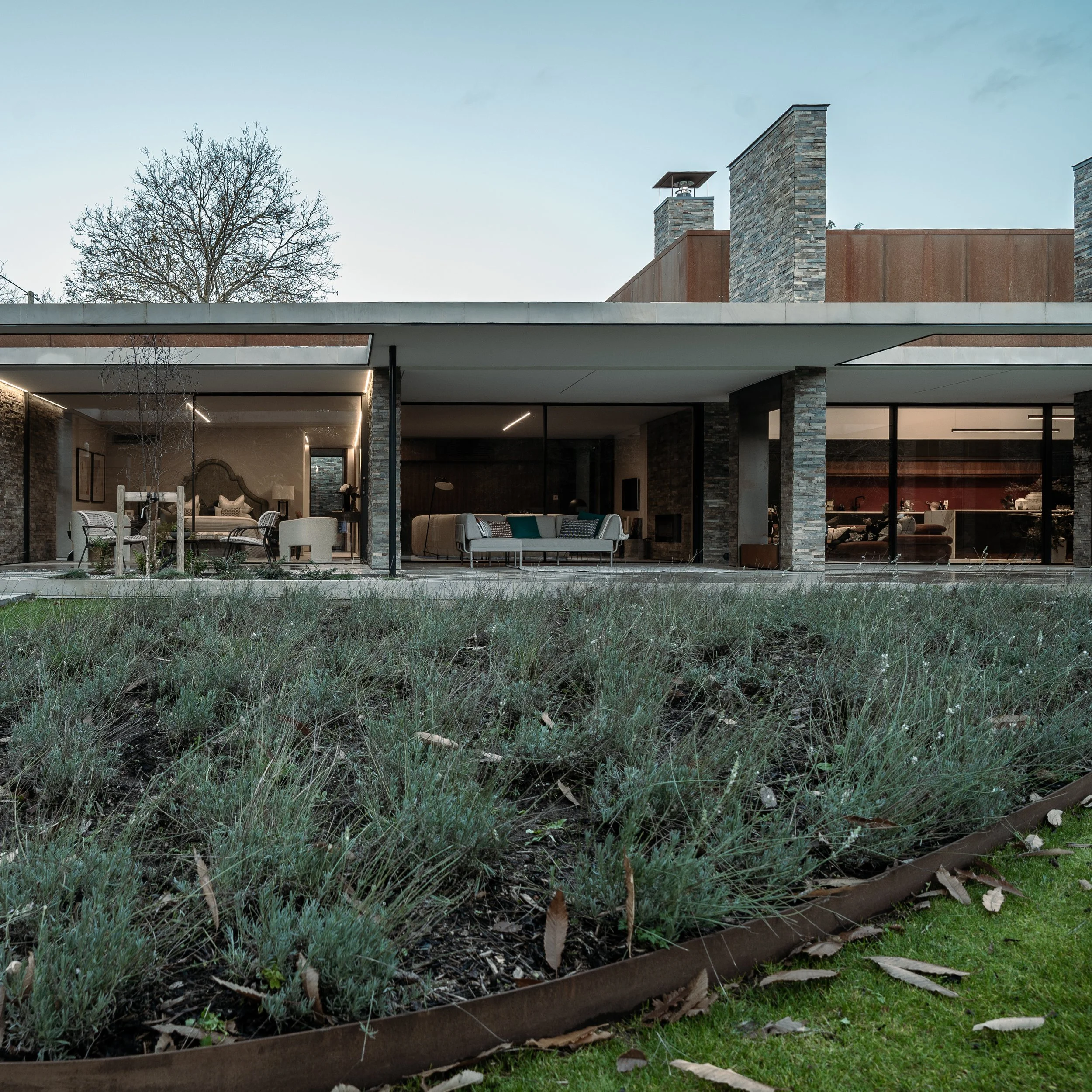 Modern house with large glass windows, stone and metal exterior, outdoor seating area, and trees in the yard during daytime.