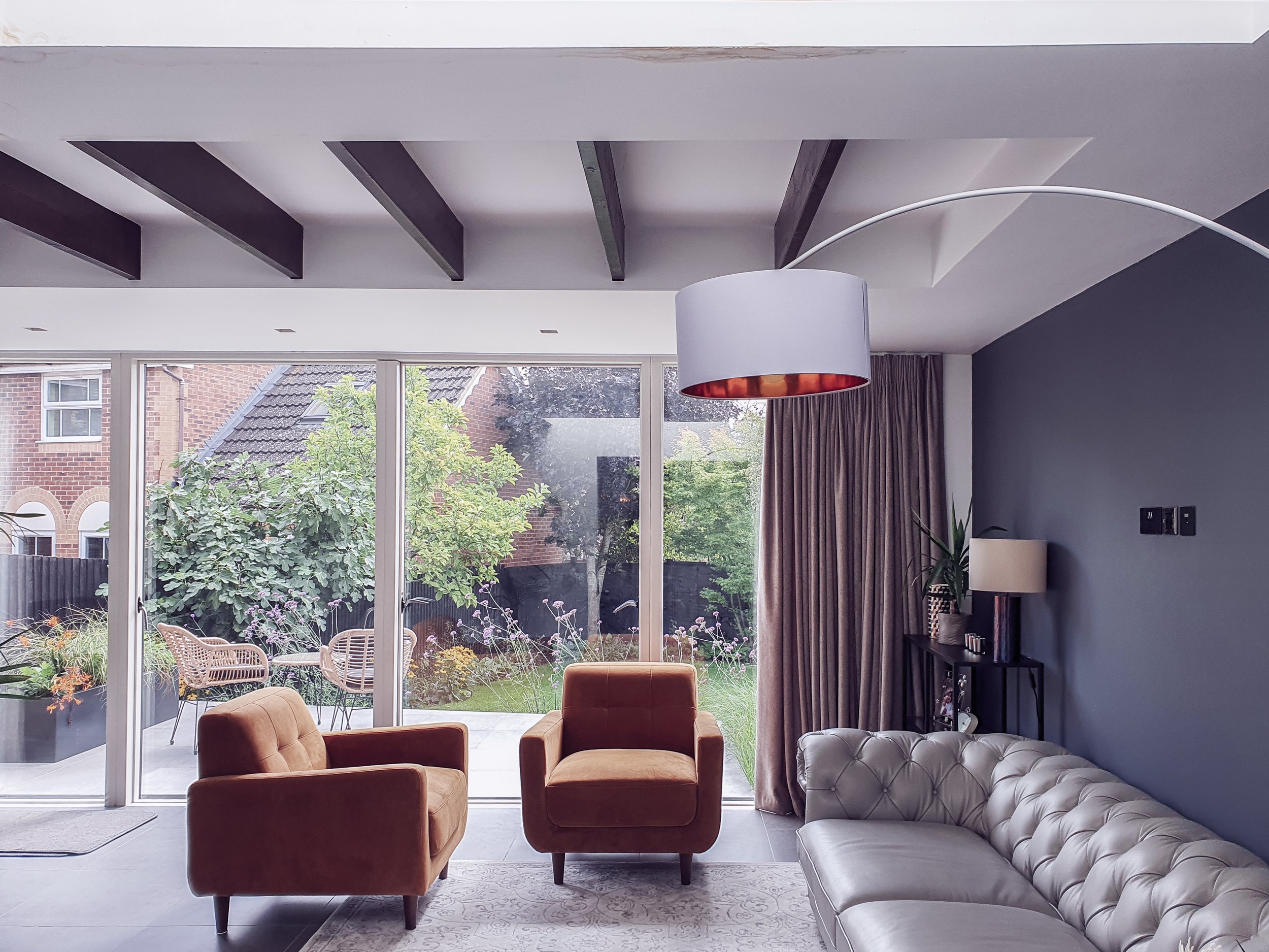 Modern living room with large glass doors opening to a garden, featuring a white and brown sectional sofa, two brown armchairs, a rug, and a ceiling lamp.