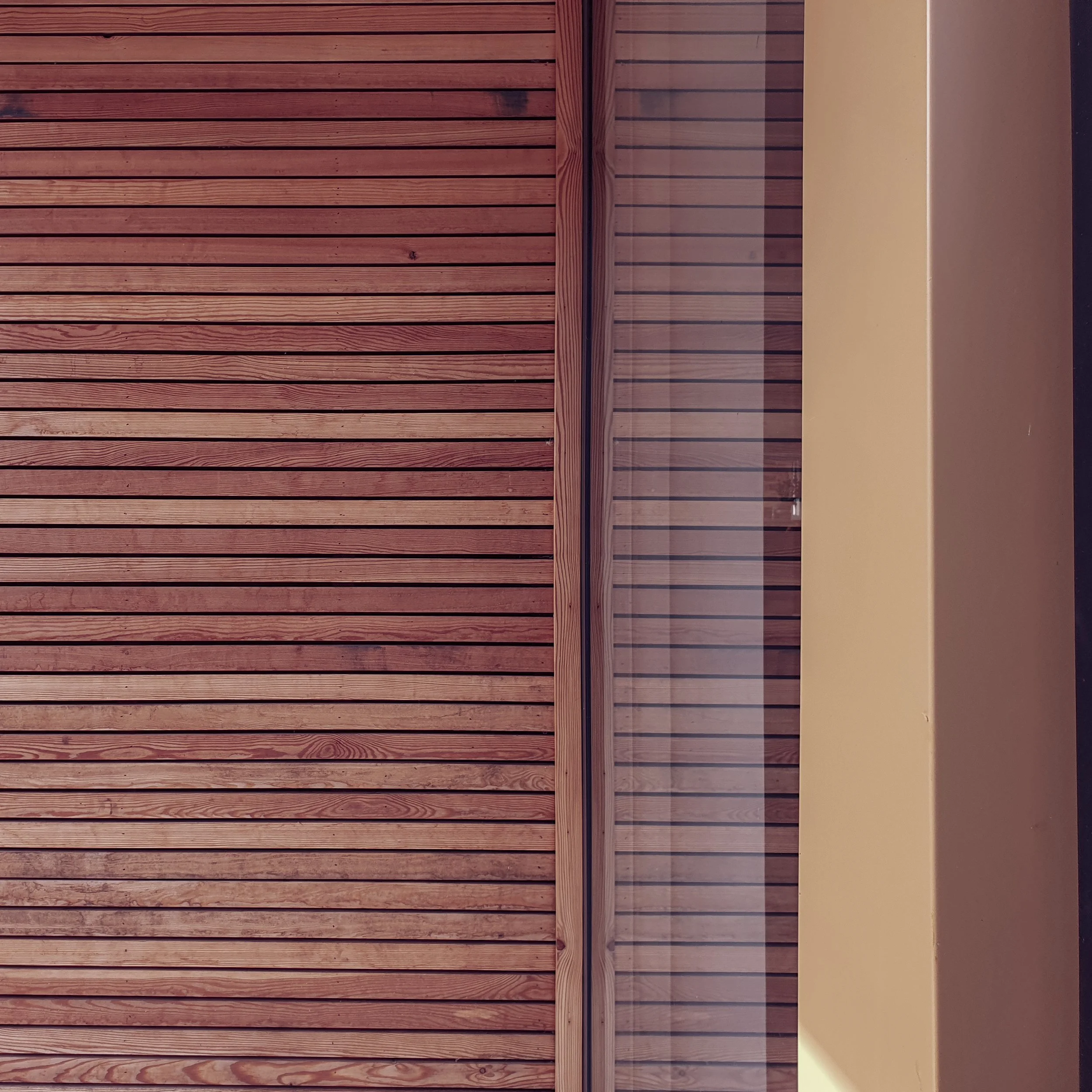 A wall of horizontal wooden planks painted in a reddish-brown color, next to a section of a beige wall with a vertical piece of wood framing a glass window or door.