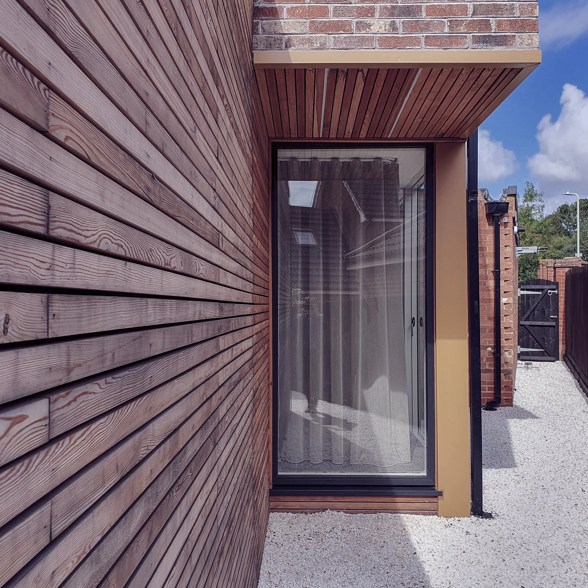 Exterior of a modern house with wooden siding, large glass door, brick wall, and cloudy sky in the background.