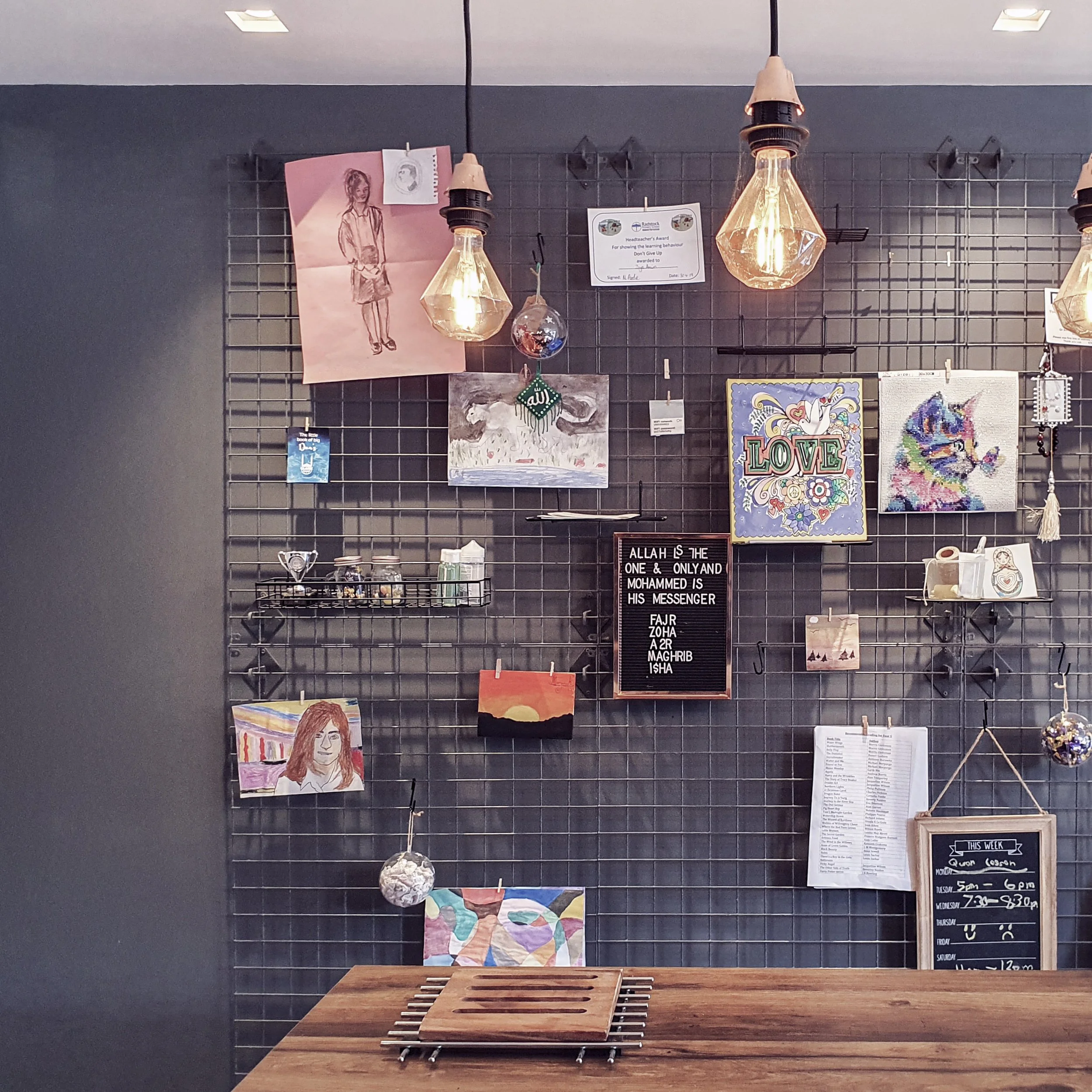 Display wall with various artwork and signs, including a pencil sketch of a girl, a color drawing of a woman, and decorative items, with a wooden table in the foreground.