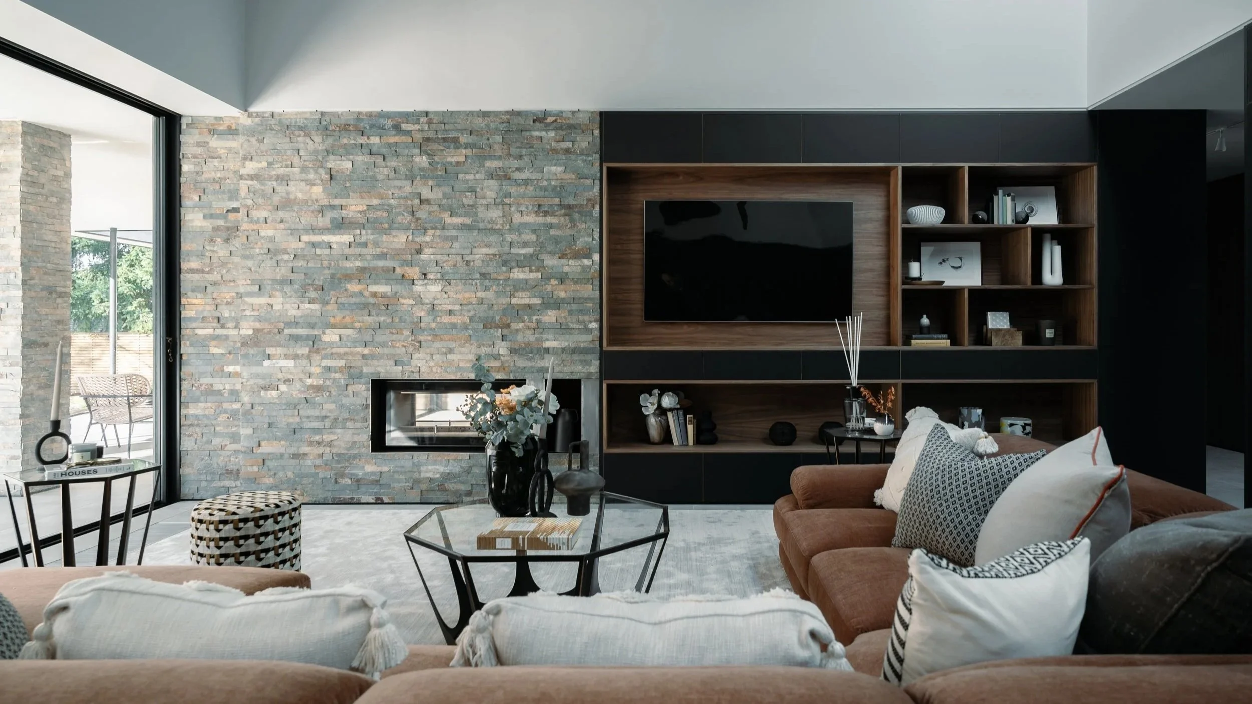 Modern living room with a stone accent wall, a wall-mounted TV, open wooden shelving with decorative items, sofa with patterned pillows, and large windows bringing in natural light.