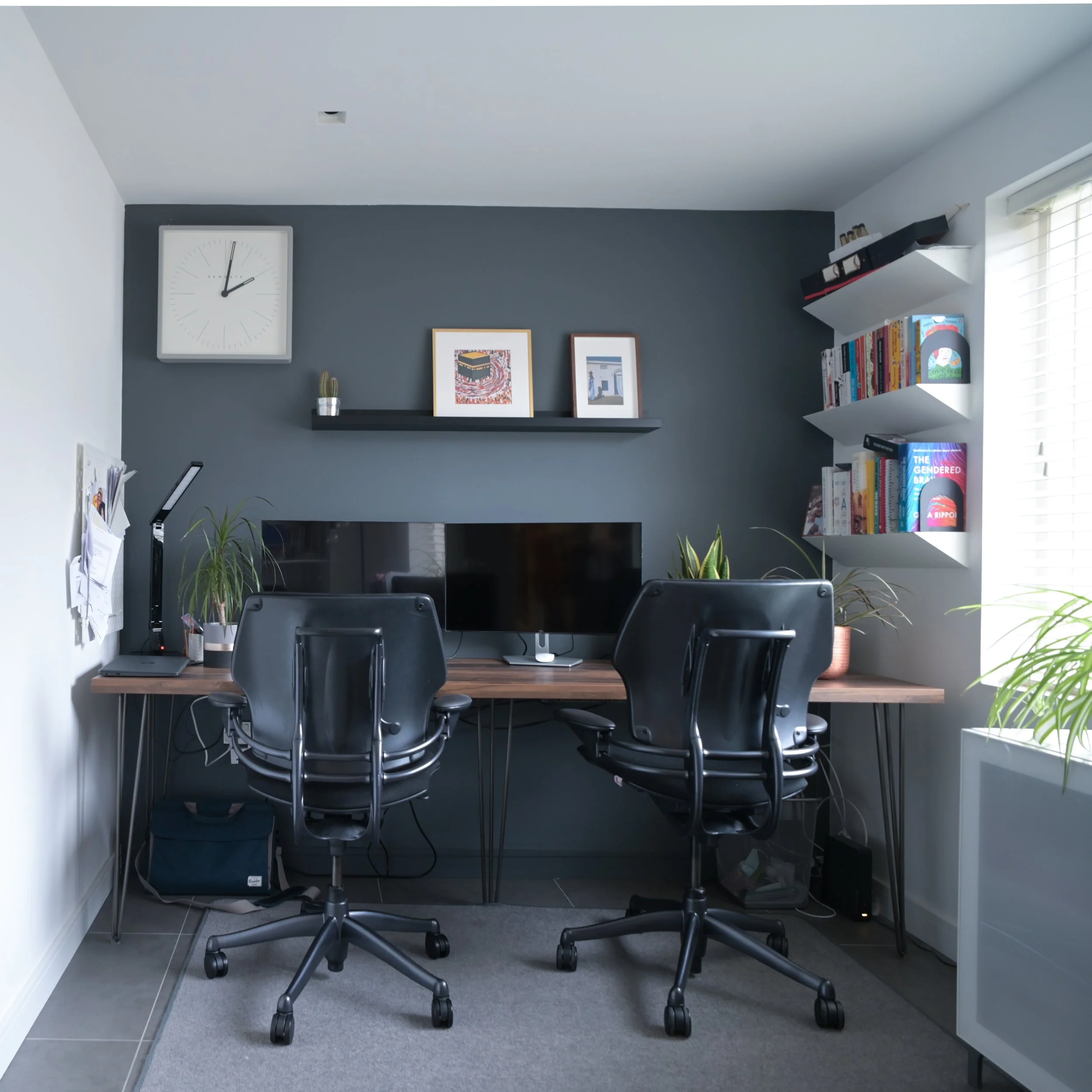 A home office with white and grey walls, a wooden desk with four black office chairs, a computer monitor, plants, books, and framed artwork, with a large wall clock showing approximately 1:09.
