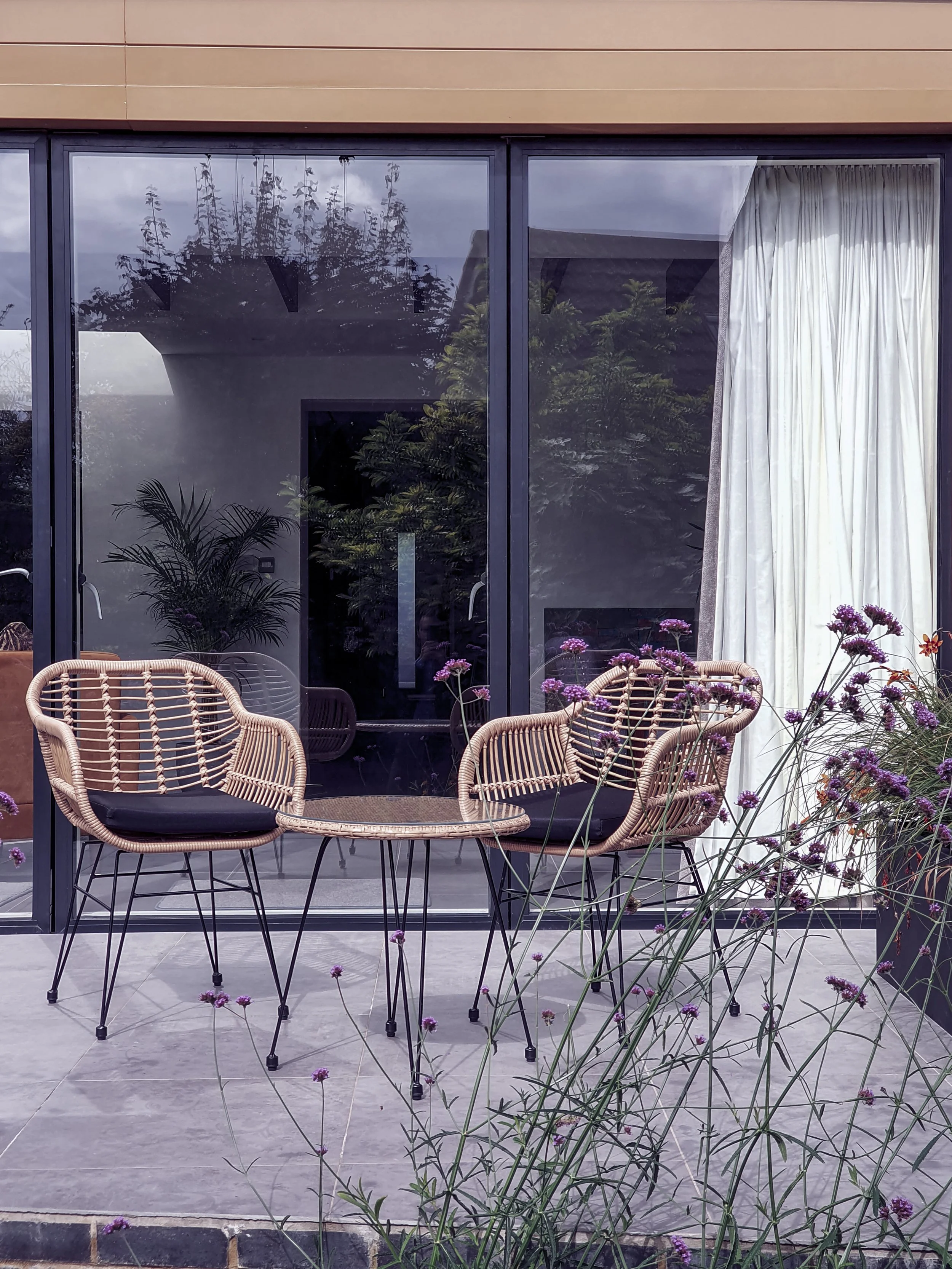 Outdoor patio area with wicker chairs and a small round table, situated in front of a glass sliding door with white curtains. Purple flowers grow in the foreground, and trees and a building are visible through the glass.