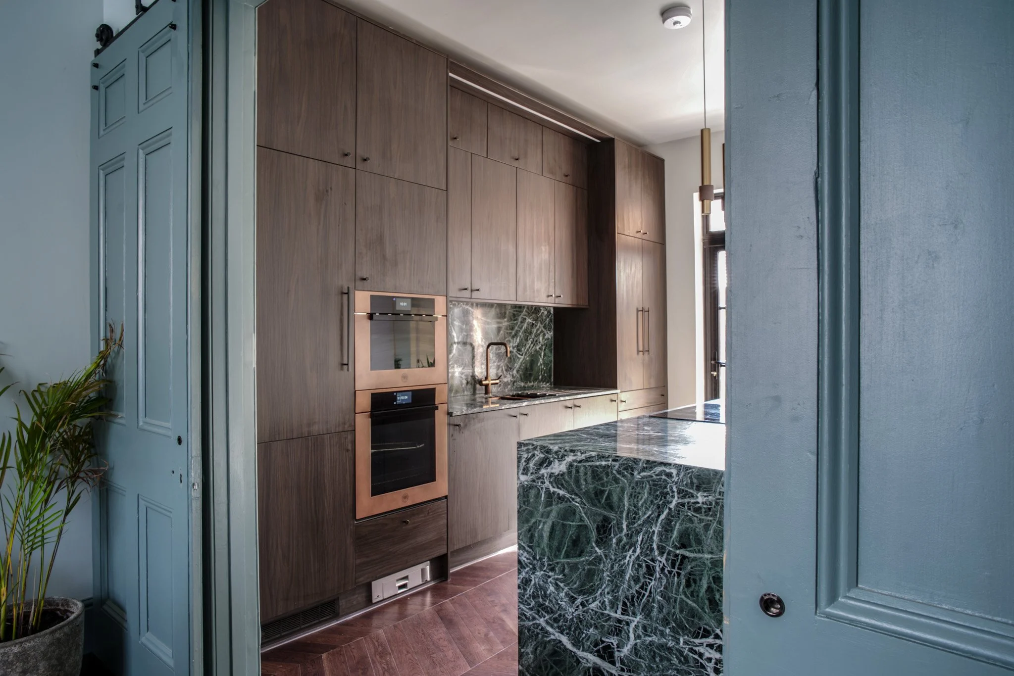 Kitchen with wooden cabinets, black marble countertops, stainless steel oven, and green marble backsplash. Part of a light blue door and potted plant are visible.