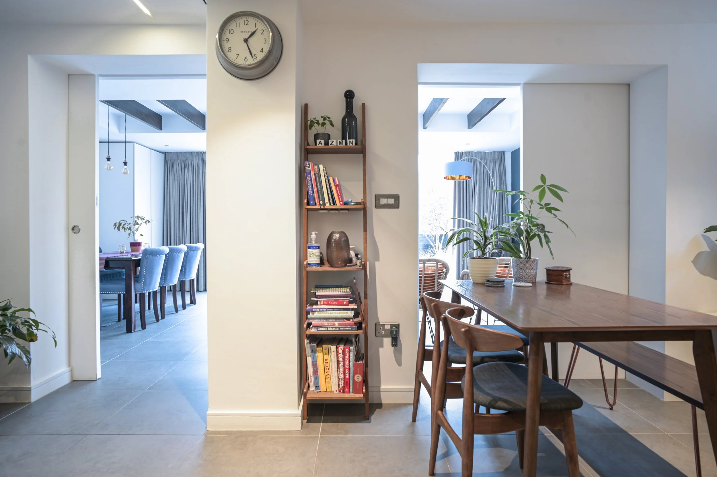 Interior view of a modern dining area with wooden chairs around a table, large potted plant, and a bookshelf against the wall. The room has large windows with curtains, and a bright, airy atmosphere with natural light.