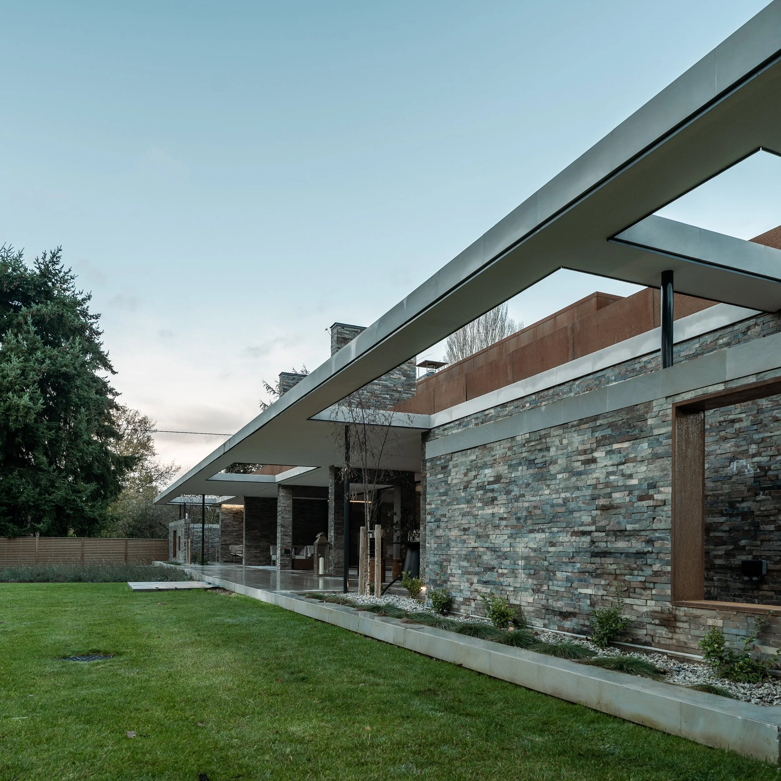 Modern building with a stone facade, large overhanging roof, and extensive outdoor greenery, including a well-maintained lawn and trees, under a clear sky.