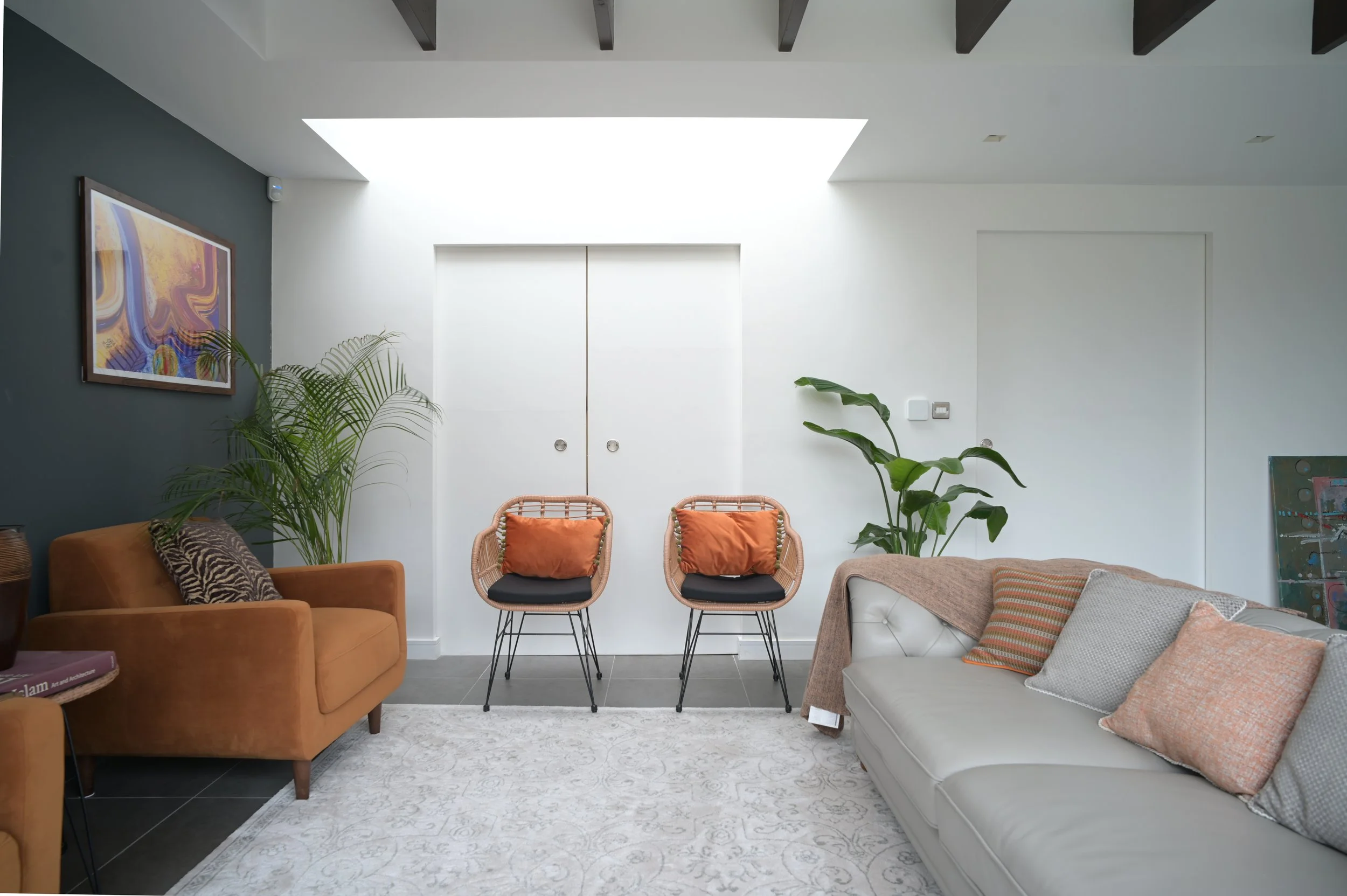 A cozy living room with a mustard-colored armchair, a white sofa with pillows, a patterned area rug, and two rattan chairs with orange cushions. There are large indoor plants, a colorful abstract painting on a green accent wall, and a white door with