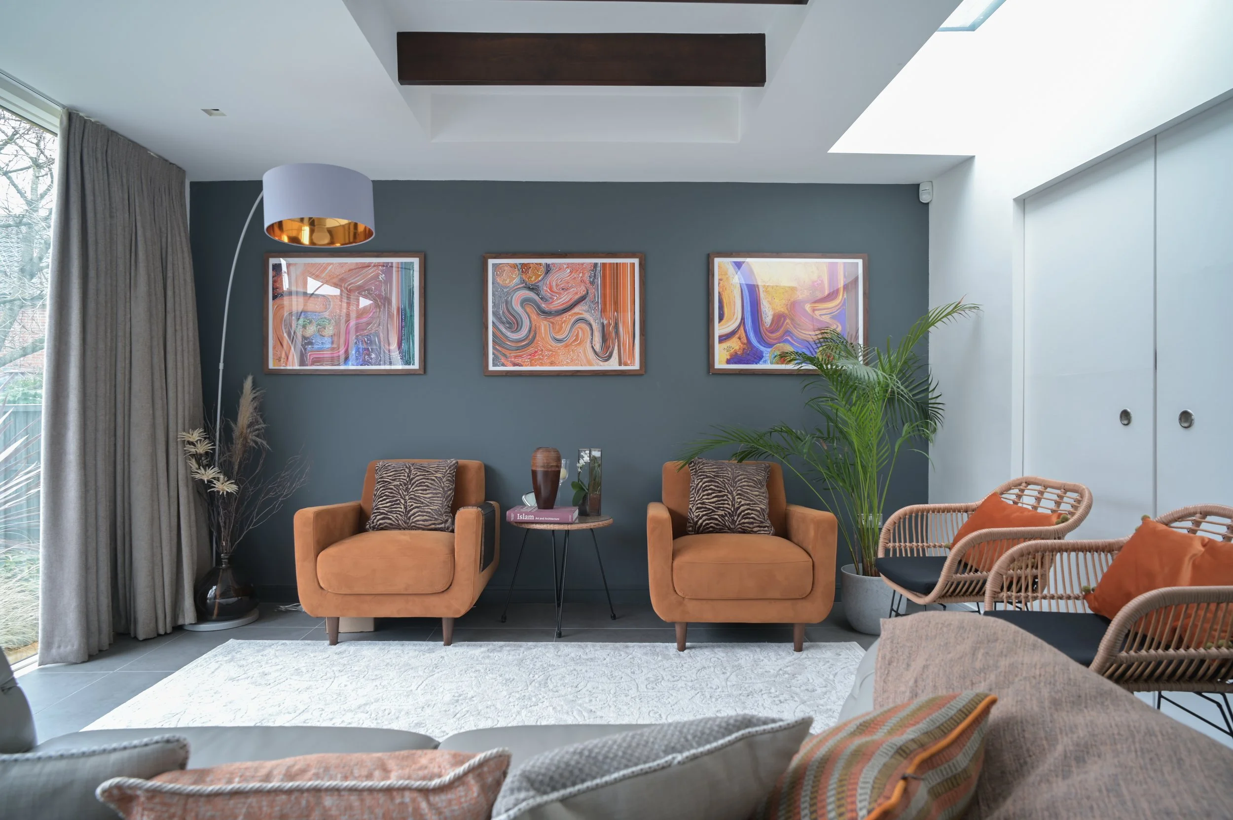 Living room with gray accent wall, three abstract artworks in wooden frames, two orange armchairs with animal print pillows, a small black side table with a book and glass, wicker chairs with orange cushions, large window with beige curtains, floor l