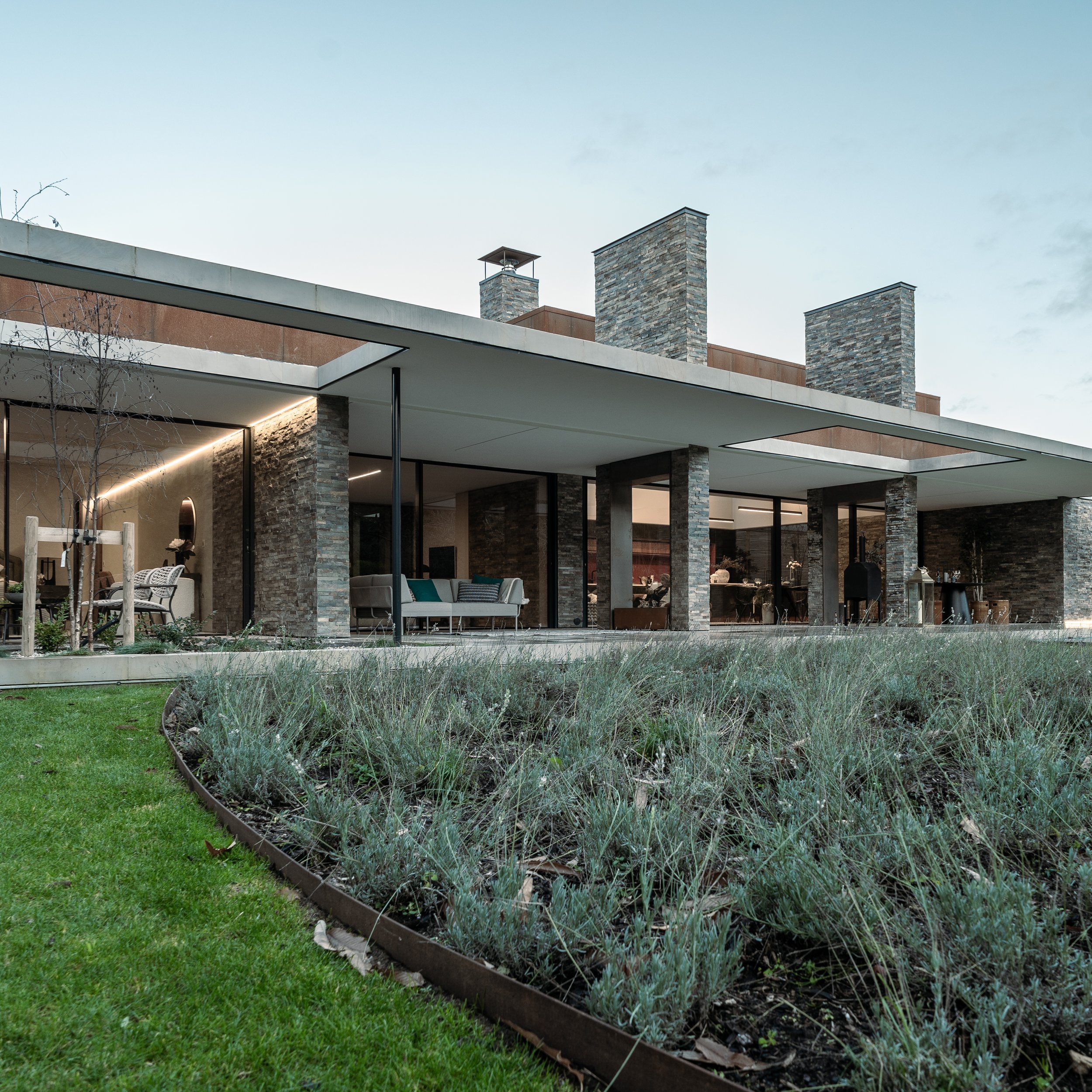 Modern house with stone chimneys, large glass windows, and outdoor seating area, under a cloudy sky.