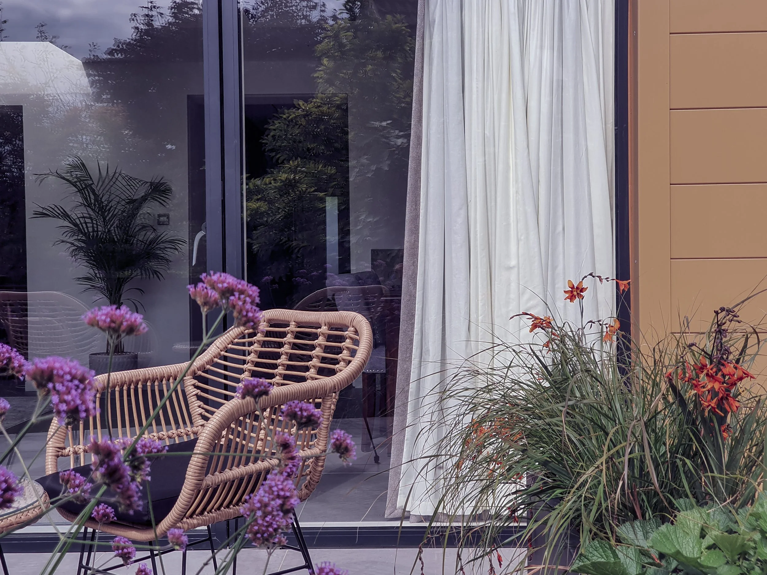 A patio with potted plants and a wicker chair in front of a large sliding glass door with white curtains. Outside, there are trees and cloudy sky visible through the glass.